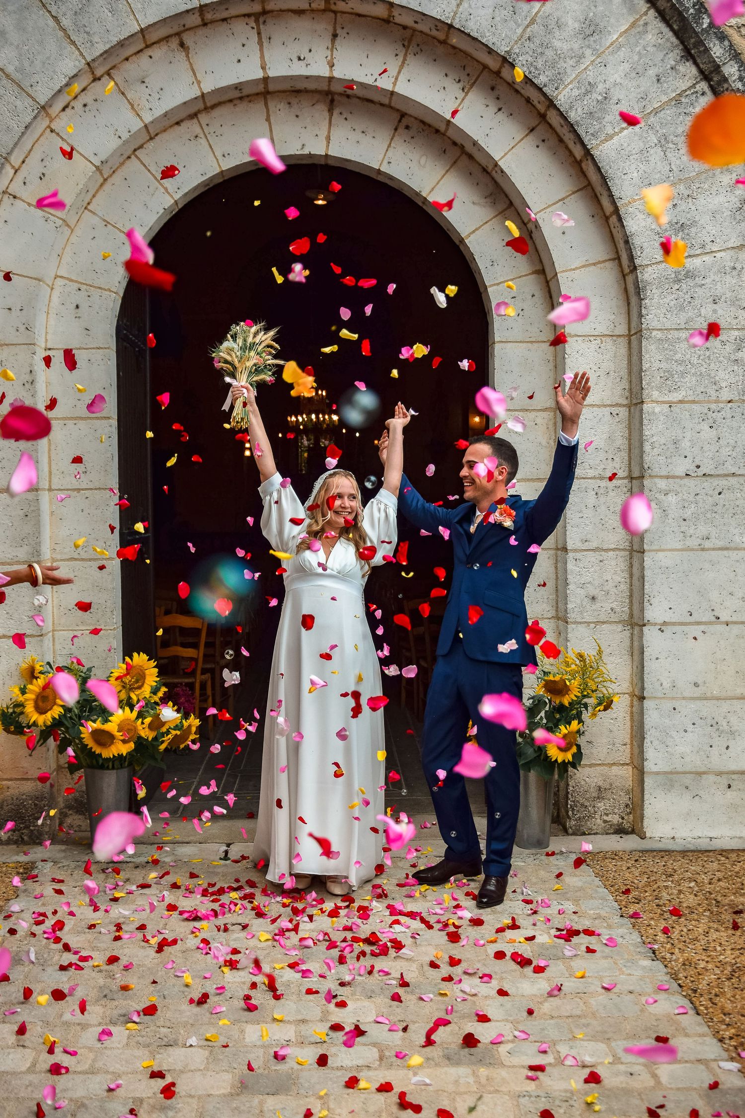 Wedding celebration as newlyweds celebrate under stone arches with pink flower petals and confetti being tossed in joy.