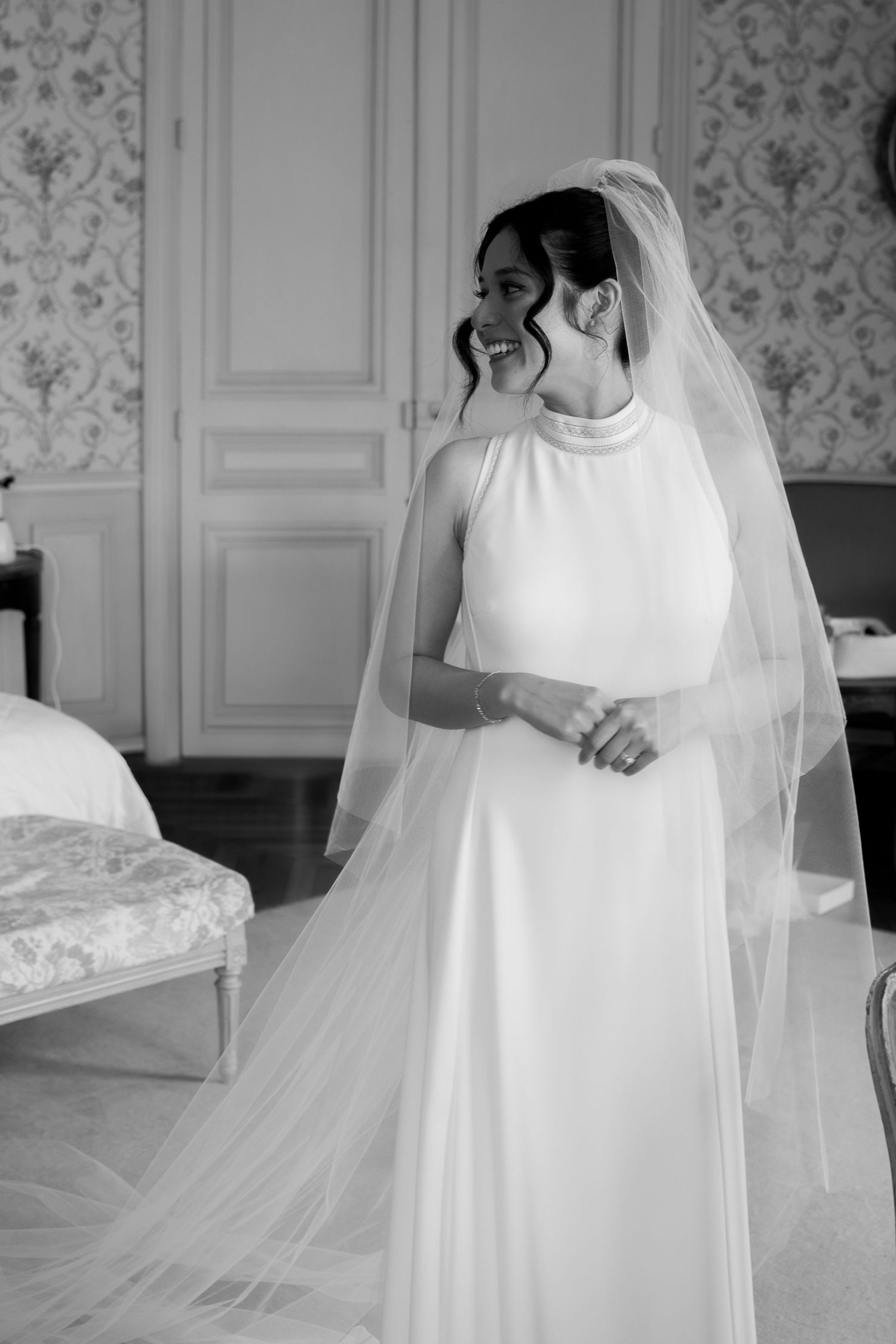 A bride stands in a white empire-waist wedding gown with mod high neckline and floor-length veil in a vintage-styled room.