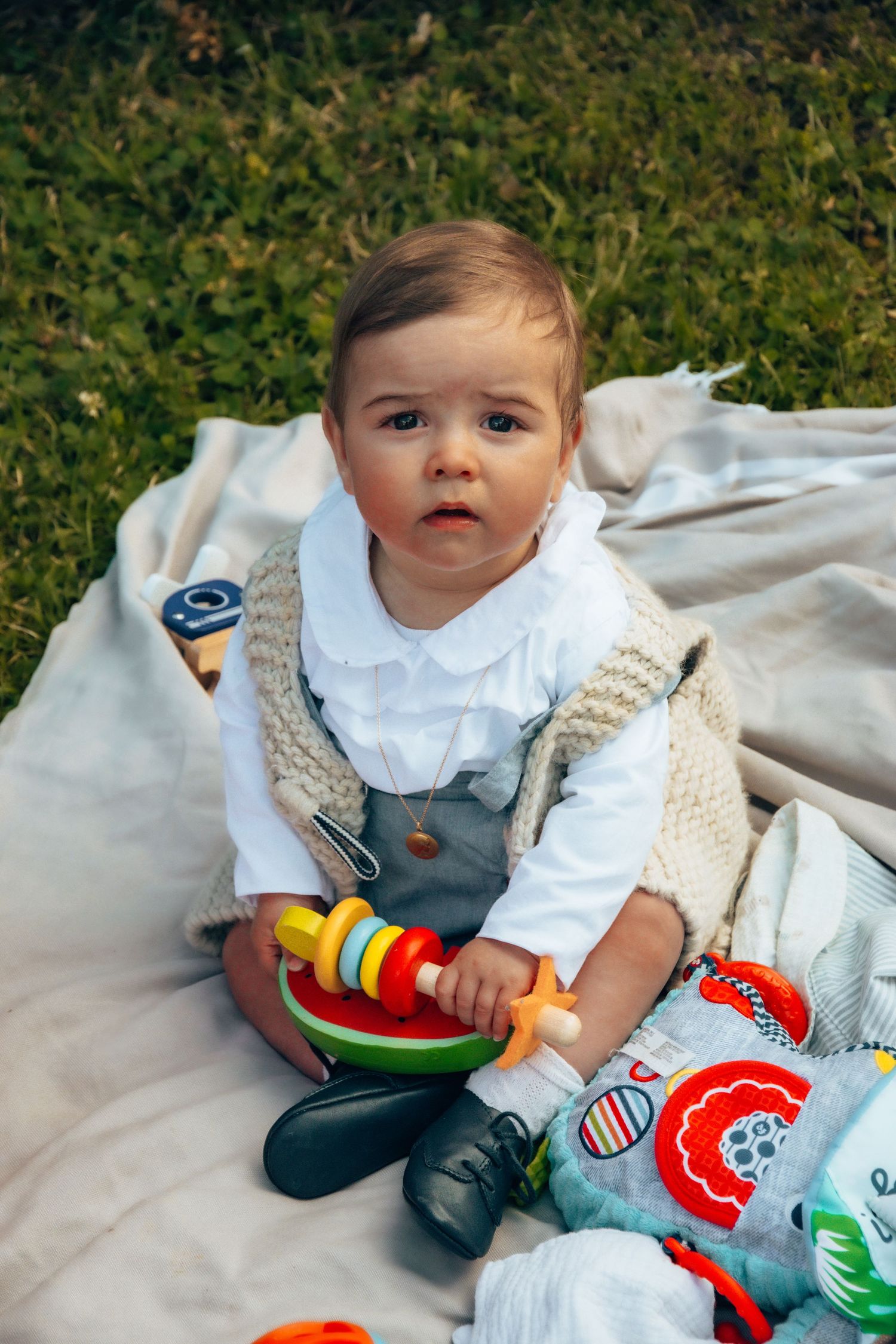 A baby in white and overalls sits on a blanket outdoors with colorful toys and plush animals.