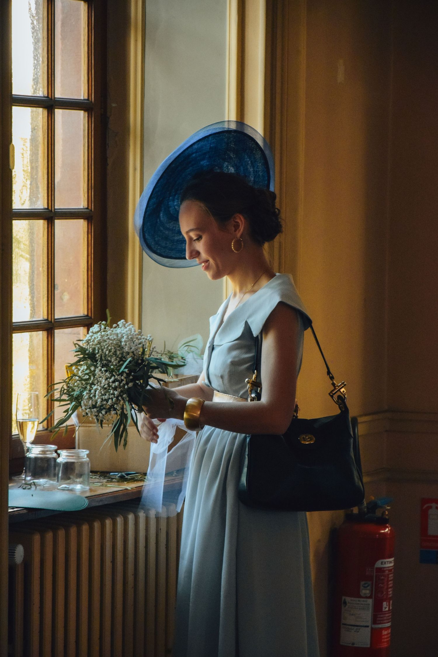 A fashionable figure in a 1950s style dress and blue hat stands by a window holding flowers with warm lighting.