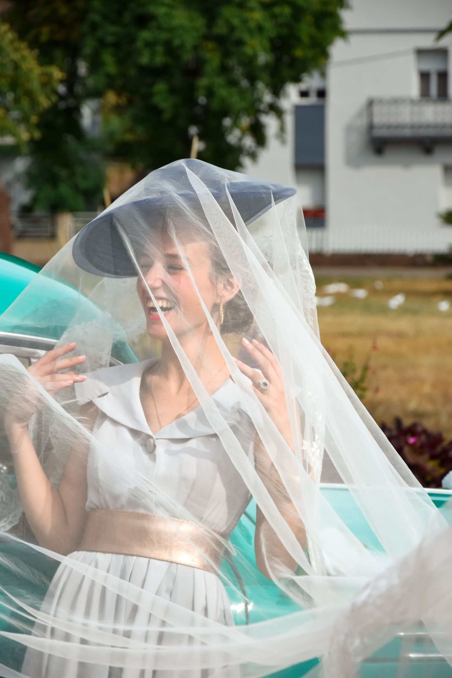 A laughing bride in a white dress covered by flowing tulle veil sits in a turquoise classic car.