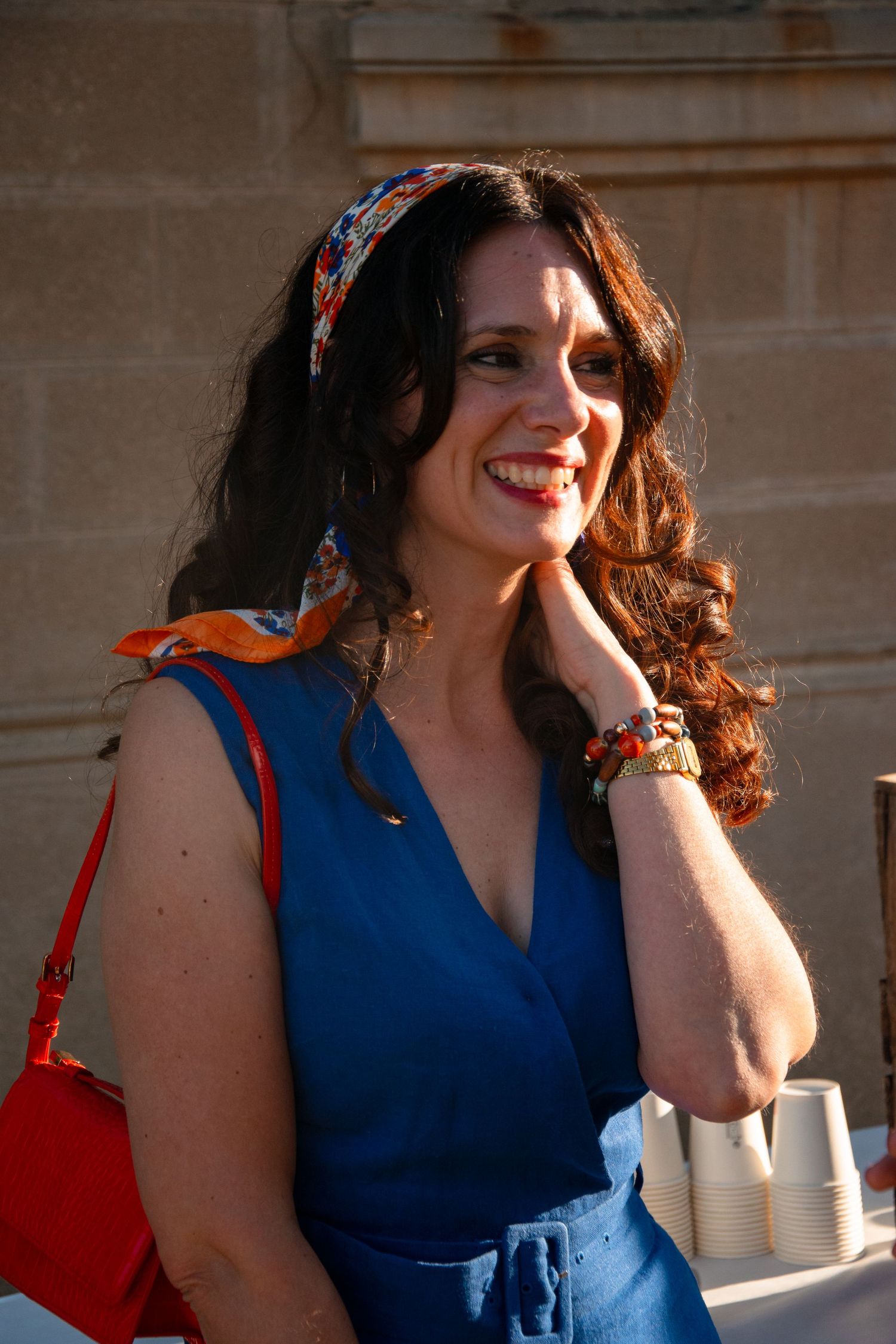 A woman in a blue dress and colorful headscarf smiling outdoors.