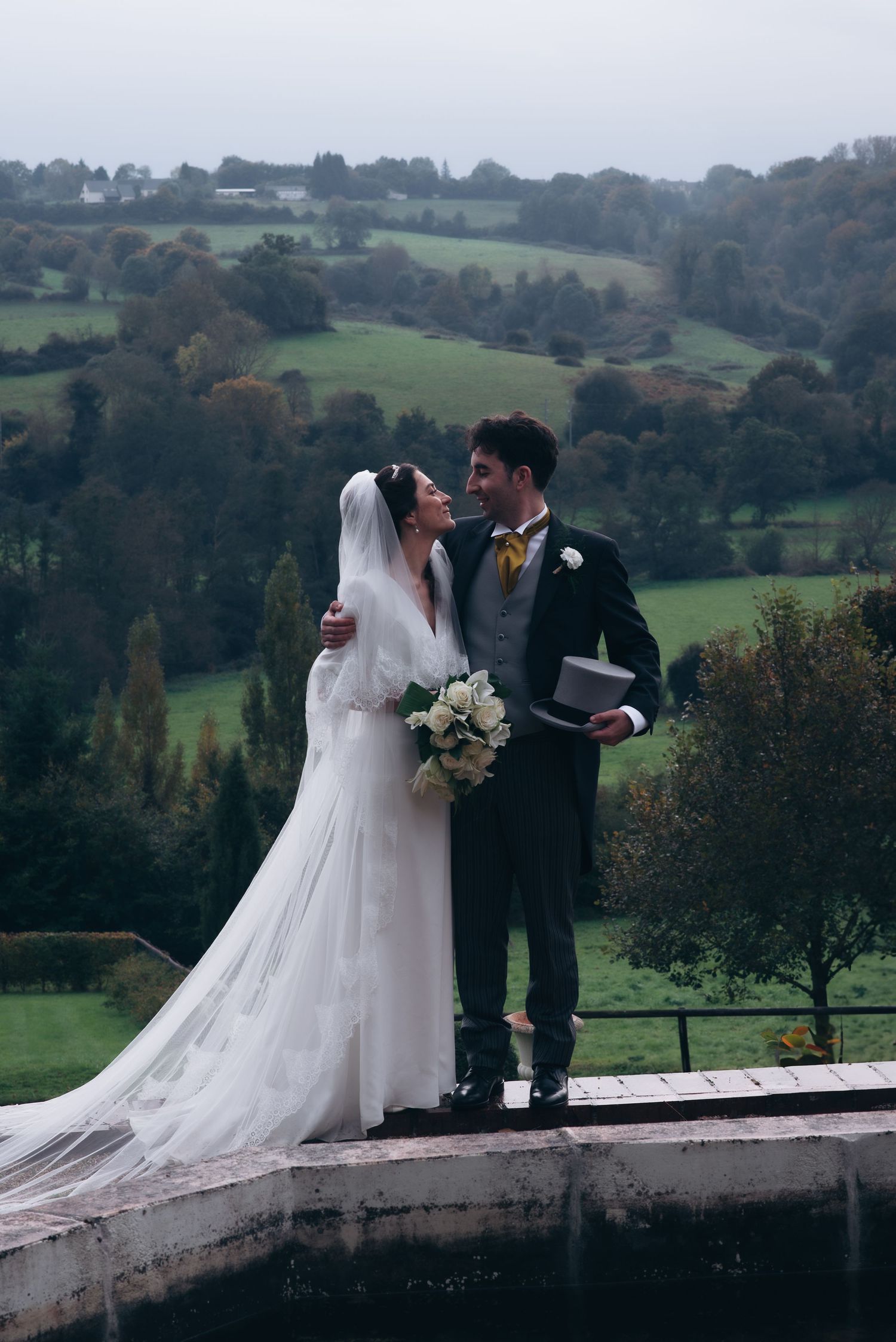 A romantic wedding photo showcasing a couple on a stone ledge overlooking rolling green countryside hills.