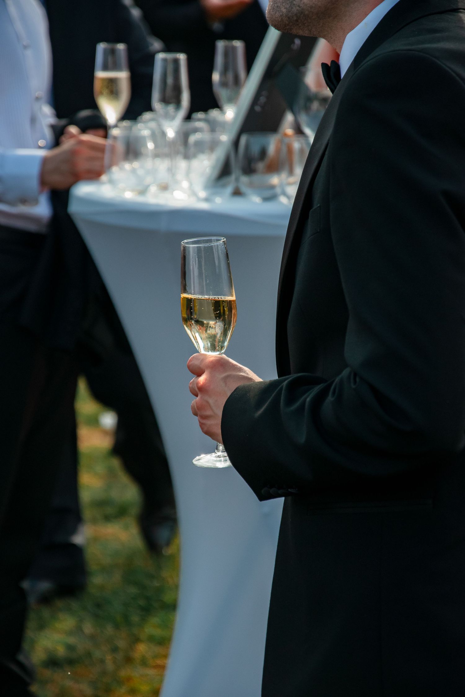 Guest in formal attire holds a champagne flute at an elegant outdoor reception with cocktail tables.