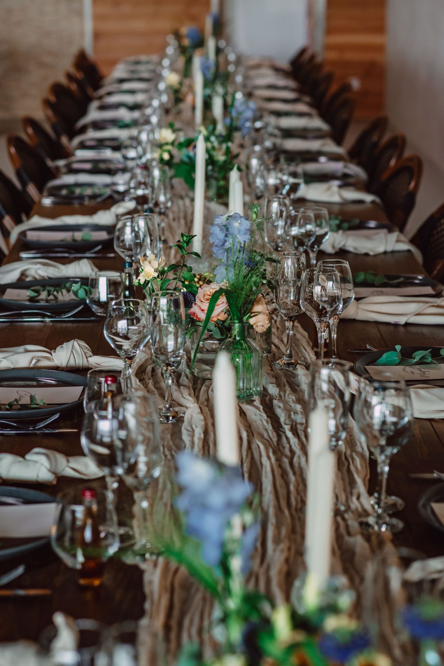 Elegant wedding reception table setting with white candles, greenery and brown runners along a rustic wooden table.
