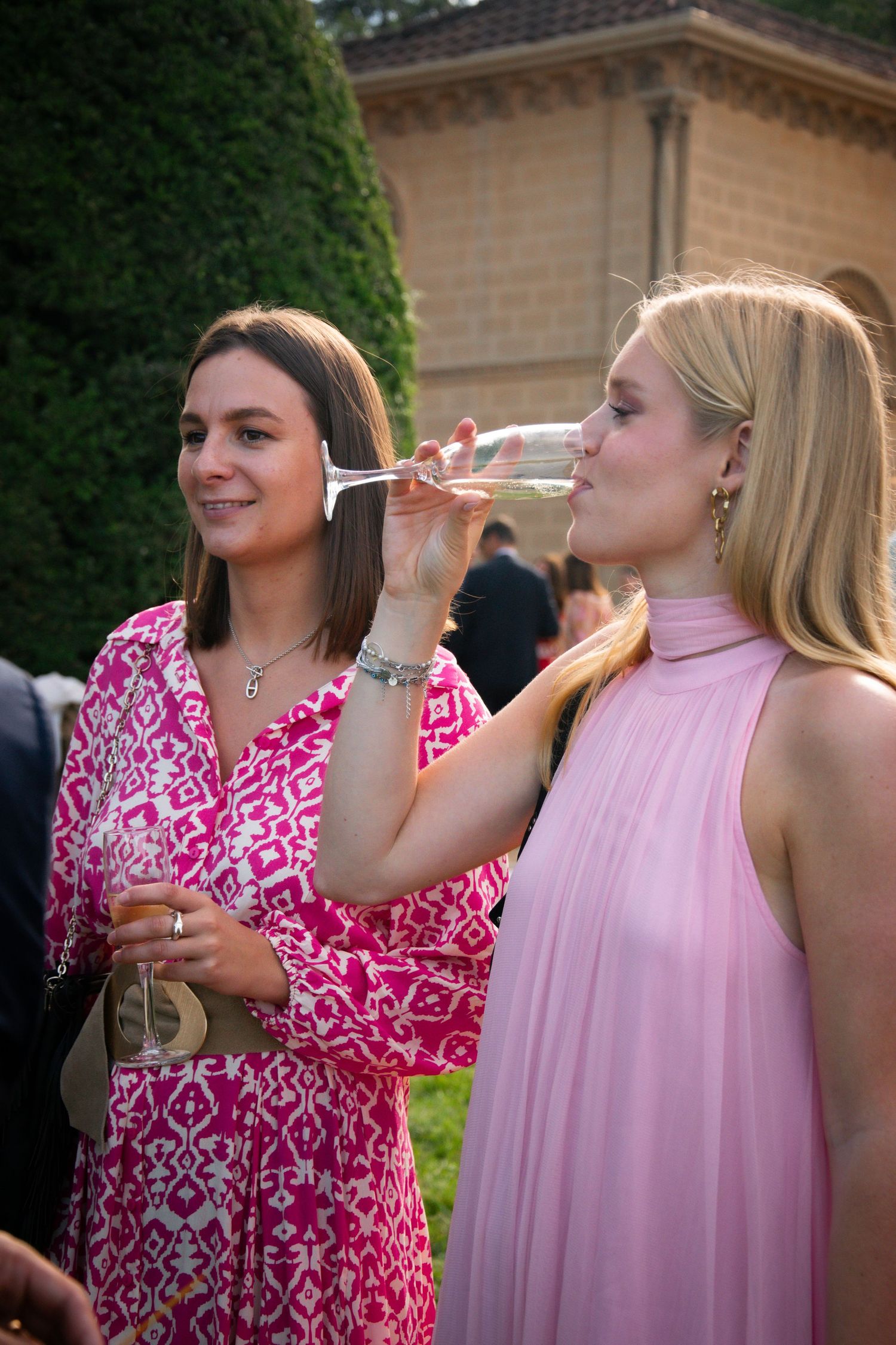 Two women in elegant dresses enjoying champagne at an outdoor event.