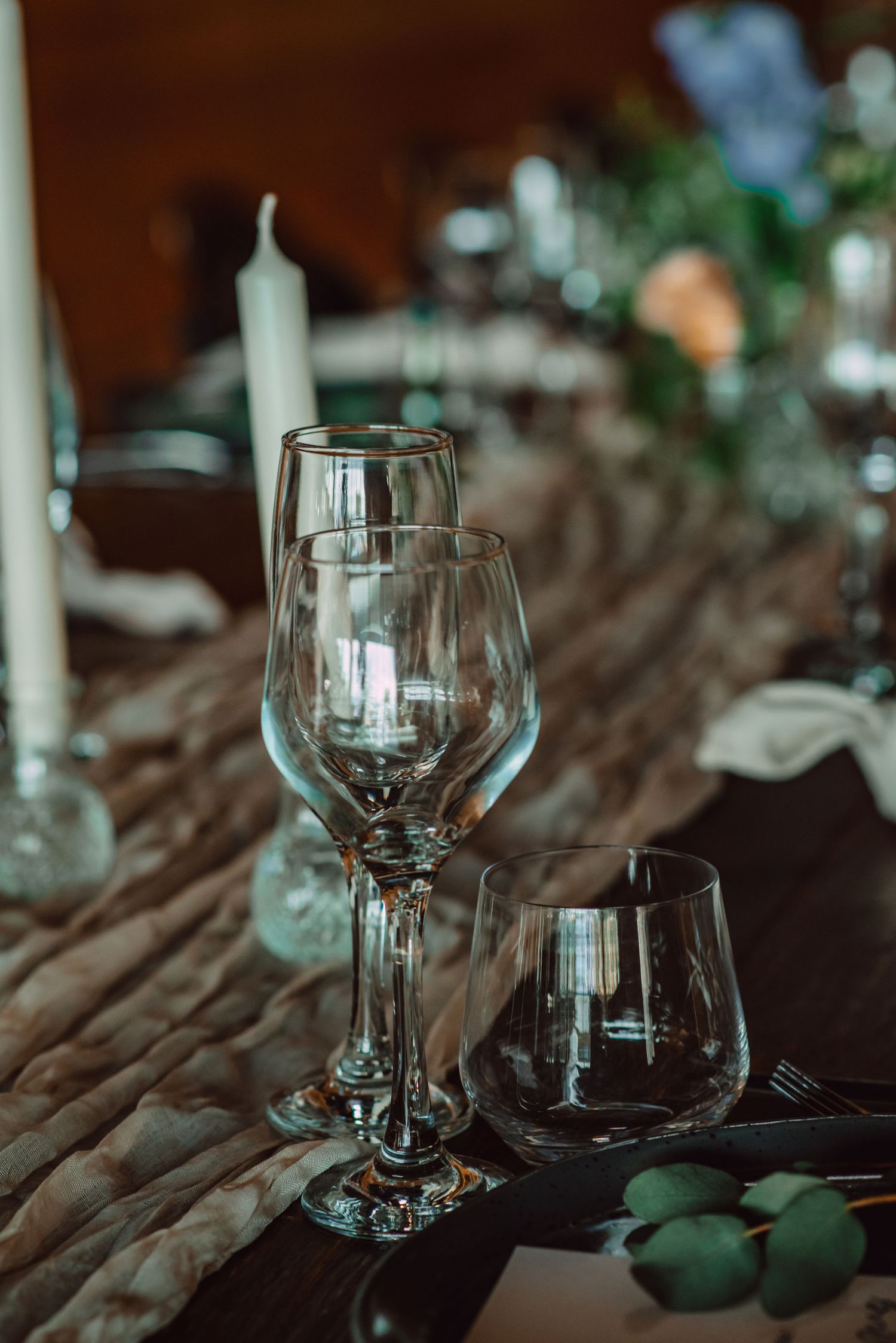 Empty wine glasses and candles set an elegant mood on a rustic wooden table with greenery accents in moody lighting.