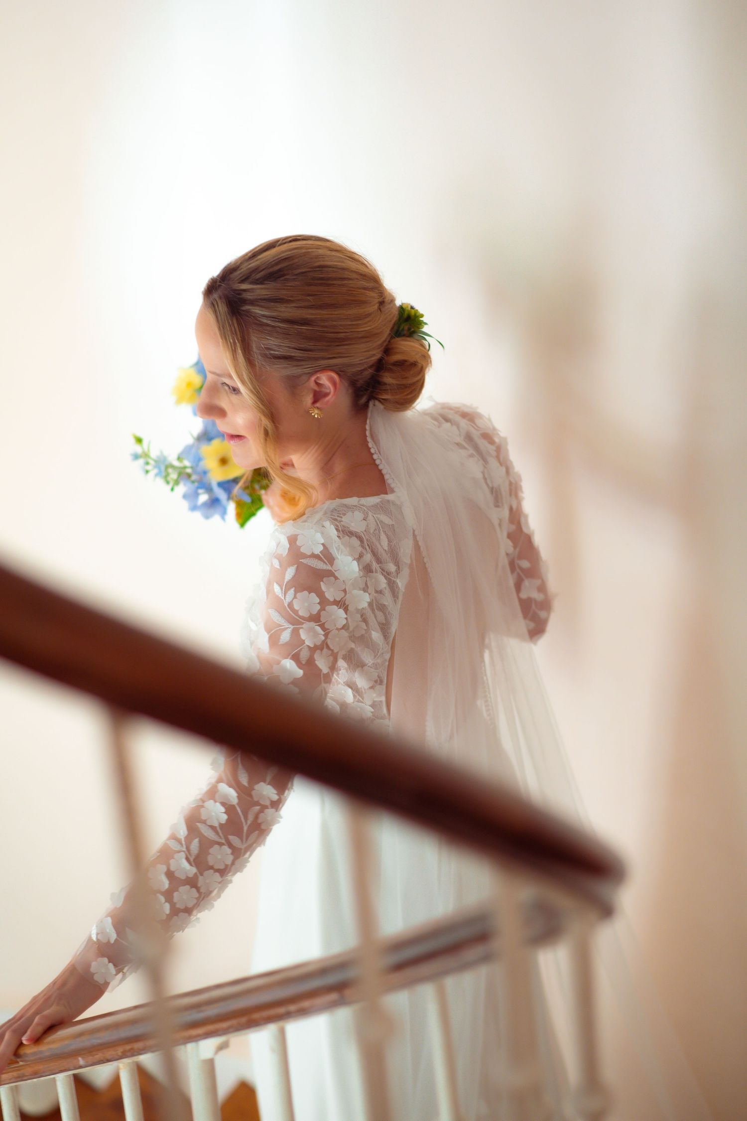 A bride in a long-sleeved lace wedding dress stands by a staircase railing holding yellow and blue flowers.
