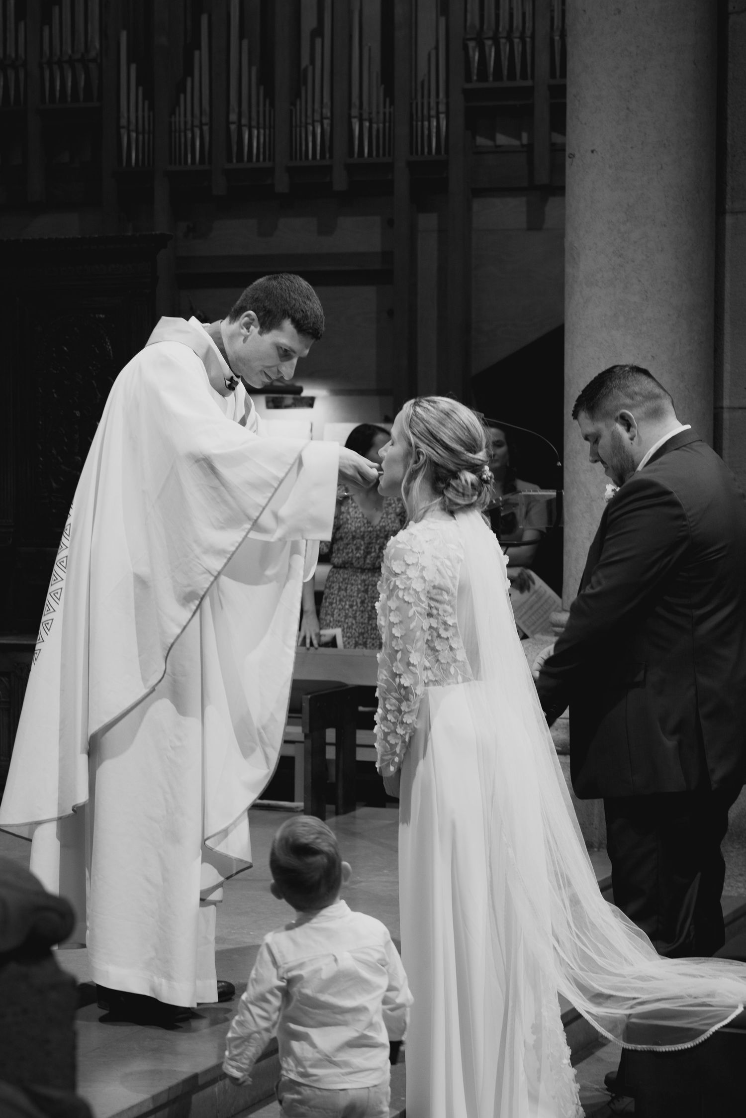 A priest in white robes blesses a couple during their wedding ceremony in a church, with a small child standing nearby.