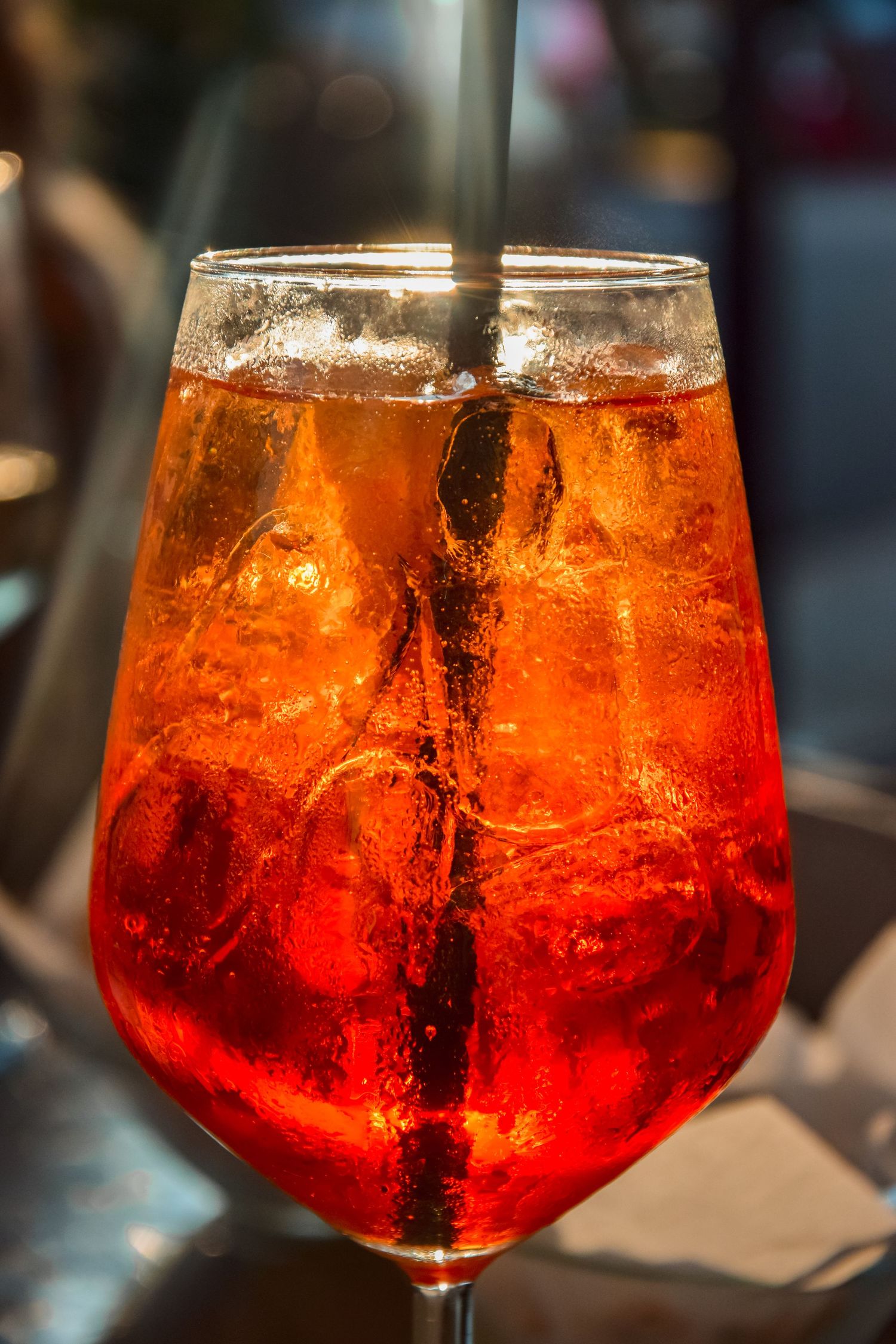 An Aperol Spritz cocktail in a wine glass filled with ice and a dark straw glows orange-red in ambient lighting.