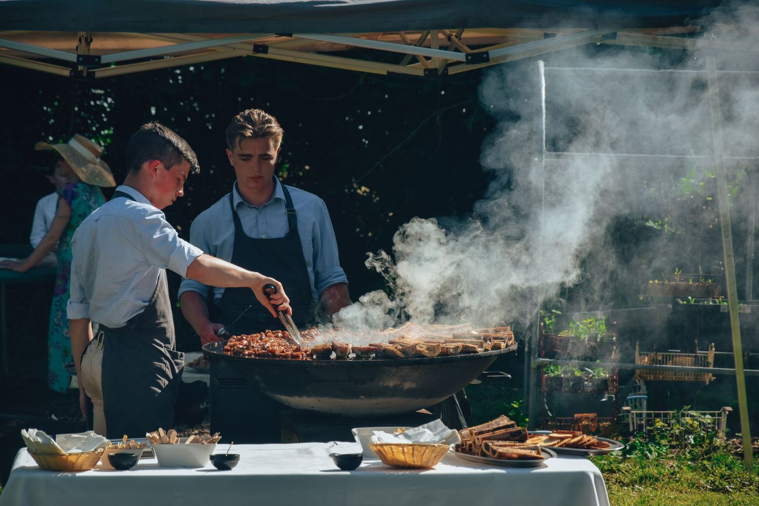 Two chefs prepare grilled meats at an outdoor buffet station with rising smoke and food platters on the table.