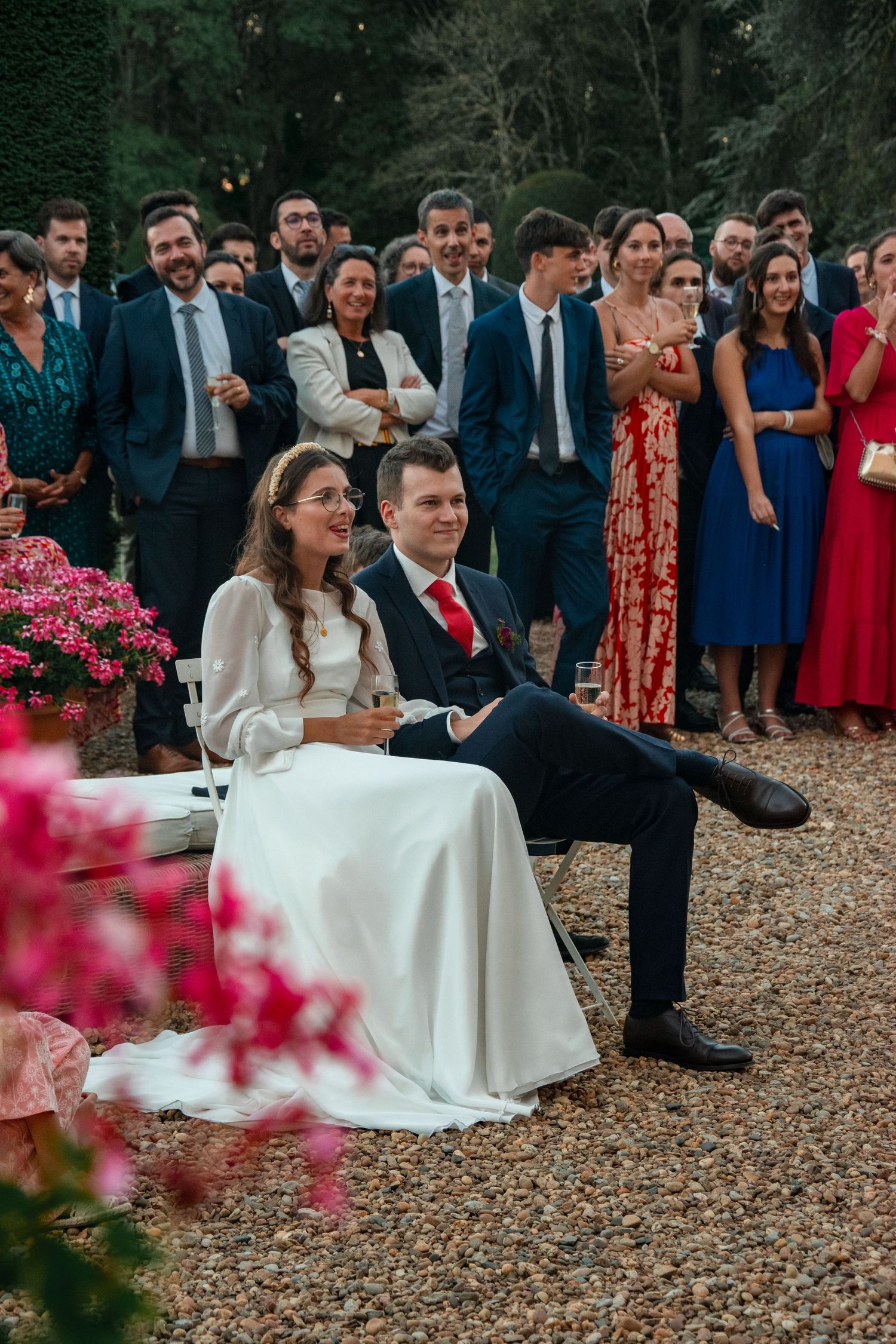 Bride and groom seated outdoors with wedding guests gathered behind pink flowers.