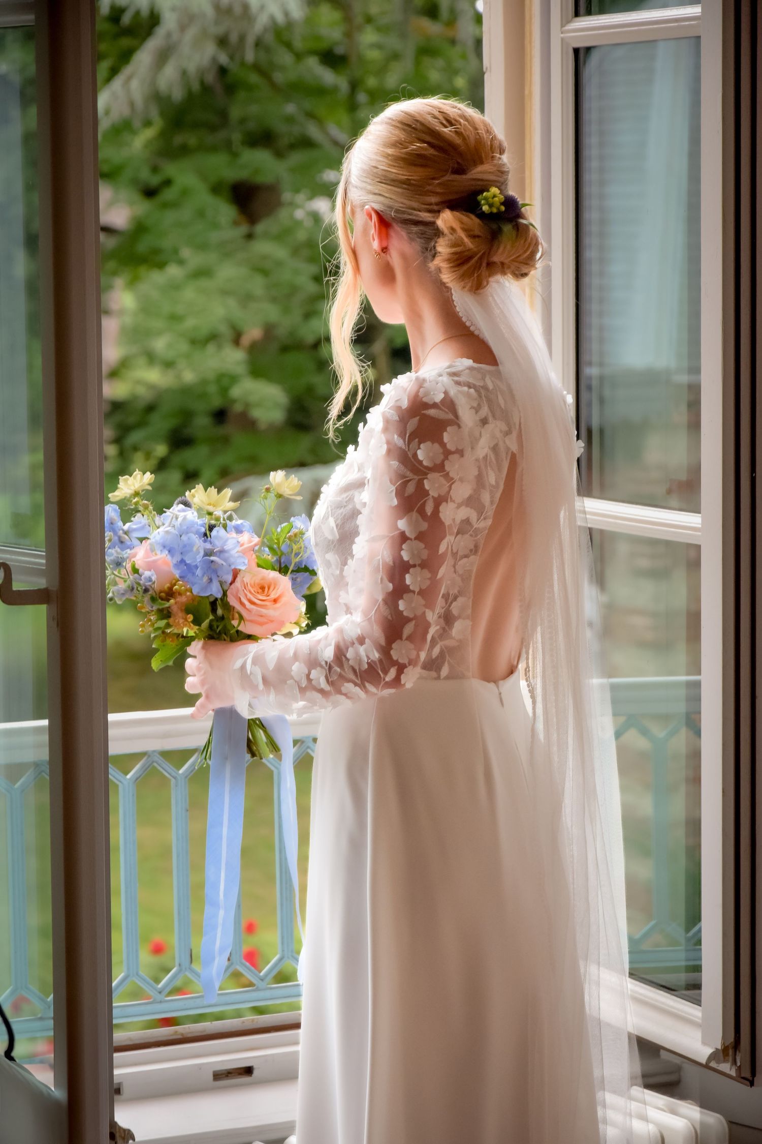 Elegant bride in lace-sleeved wedding dress with open back holds colorful bouquet while looking out balcony window.