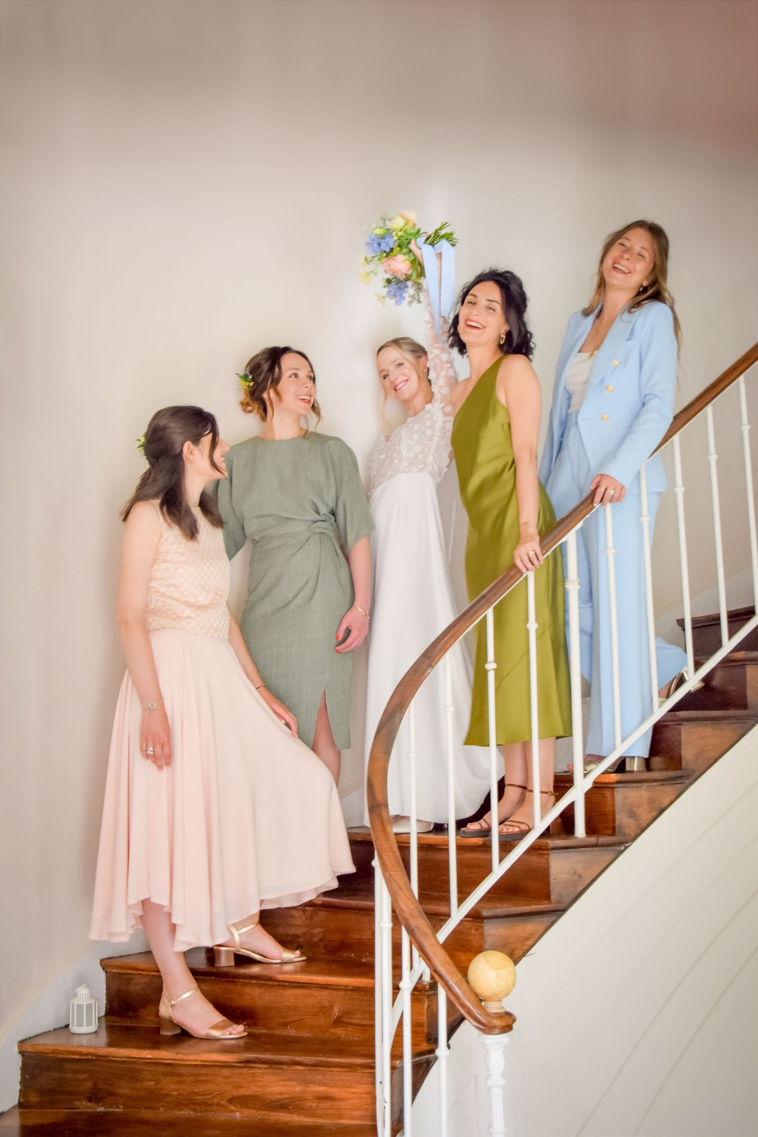 A group of women in pastel dresses stand and laugh together on a curved wooden staircase with white railing.