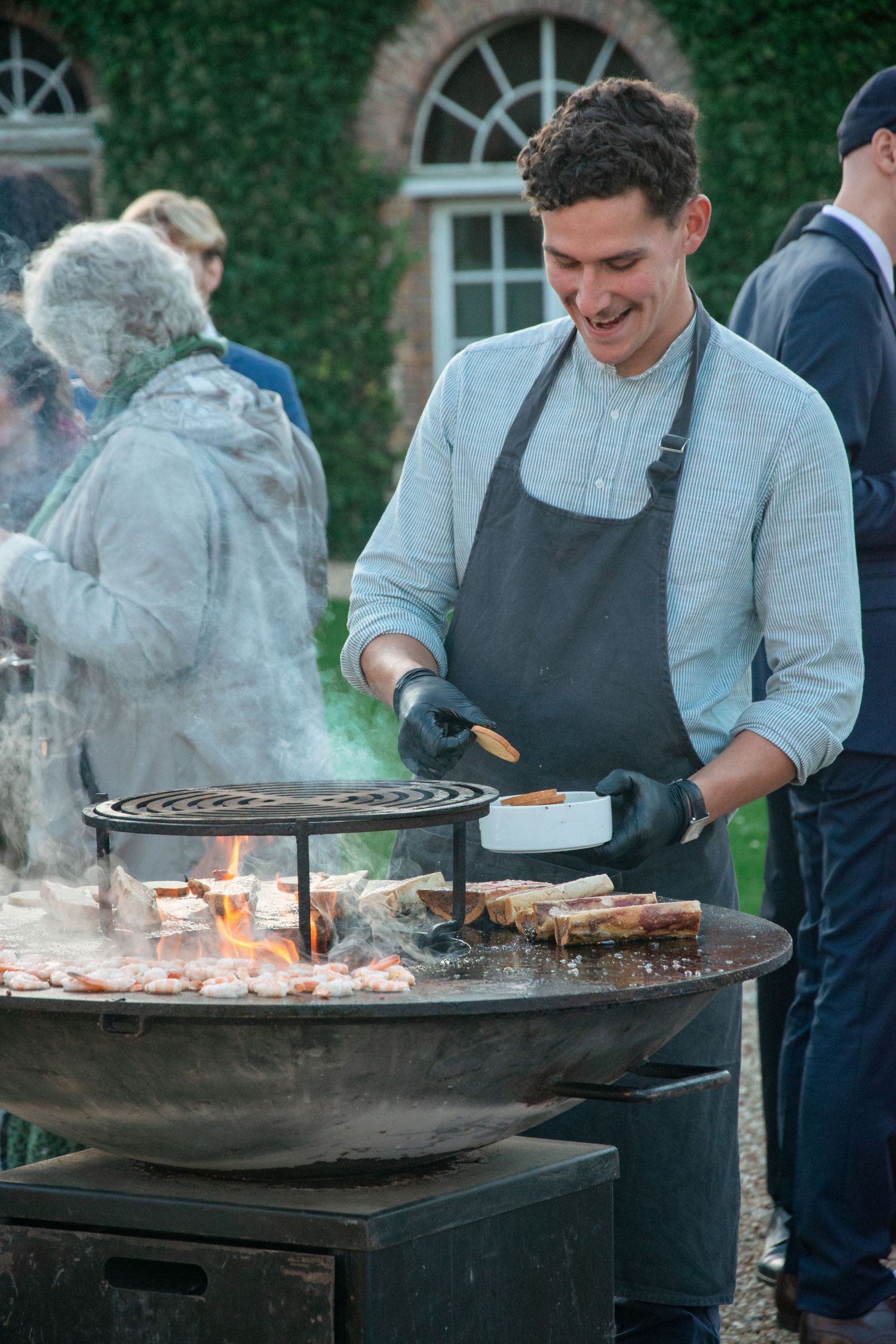 Chef grilling food on an outdoor barbecue with smoke rising from the grill at an elegant venue.