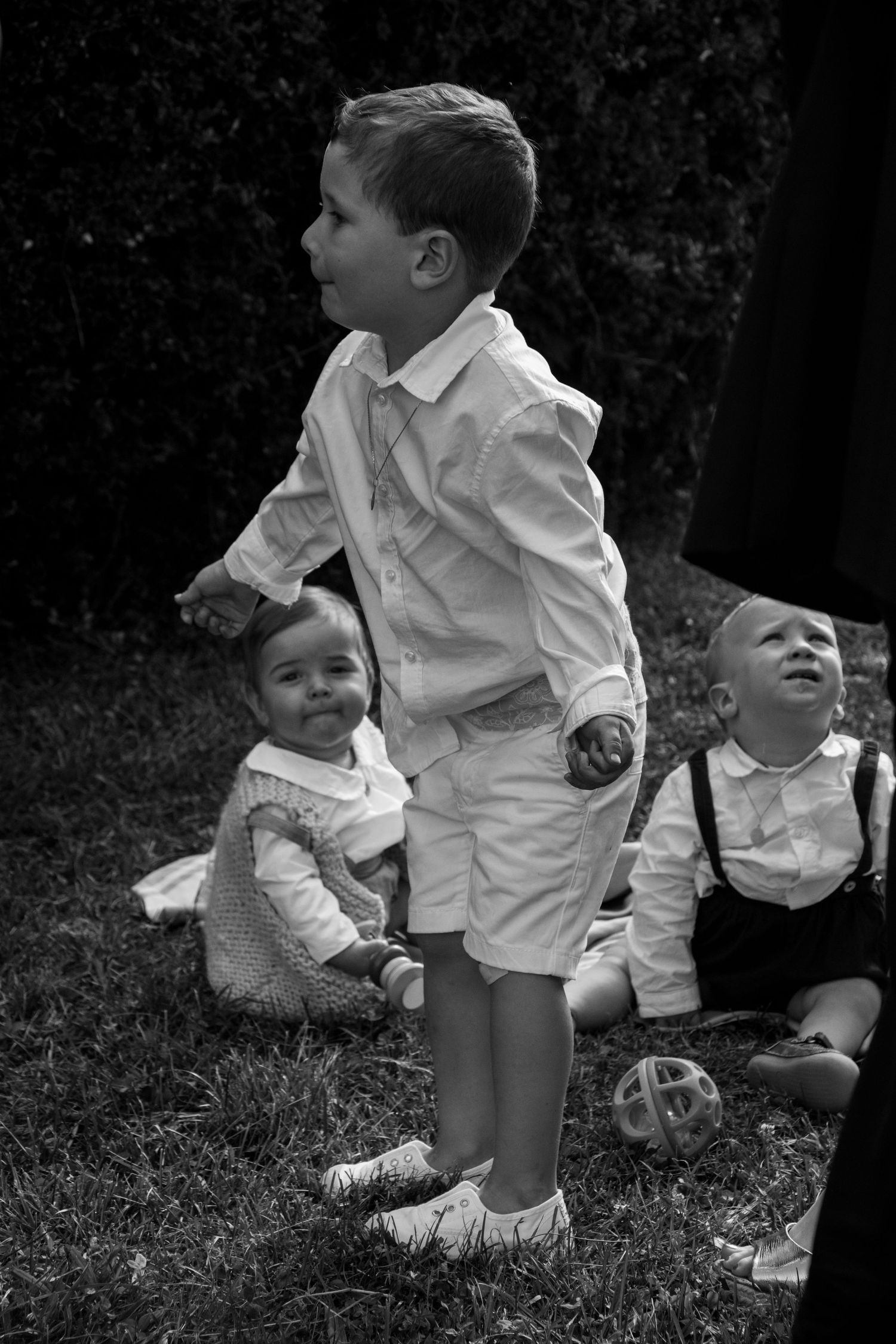 Black and white photograph of three young children in formal attire sitting and standing outdoors on grass.