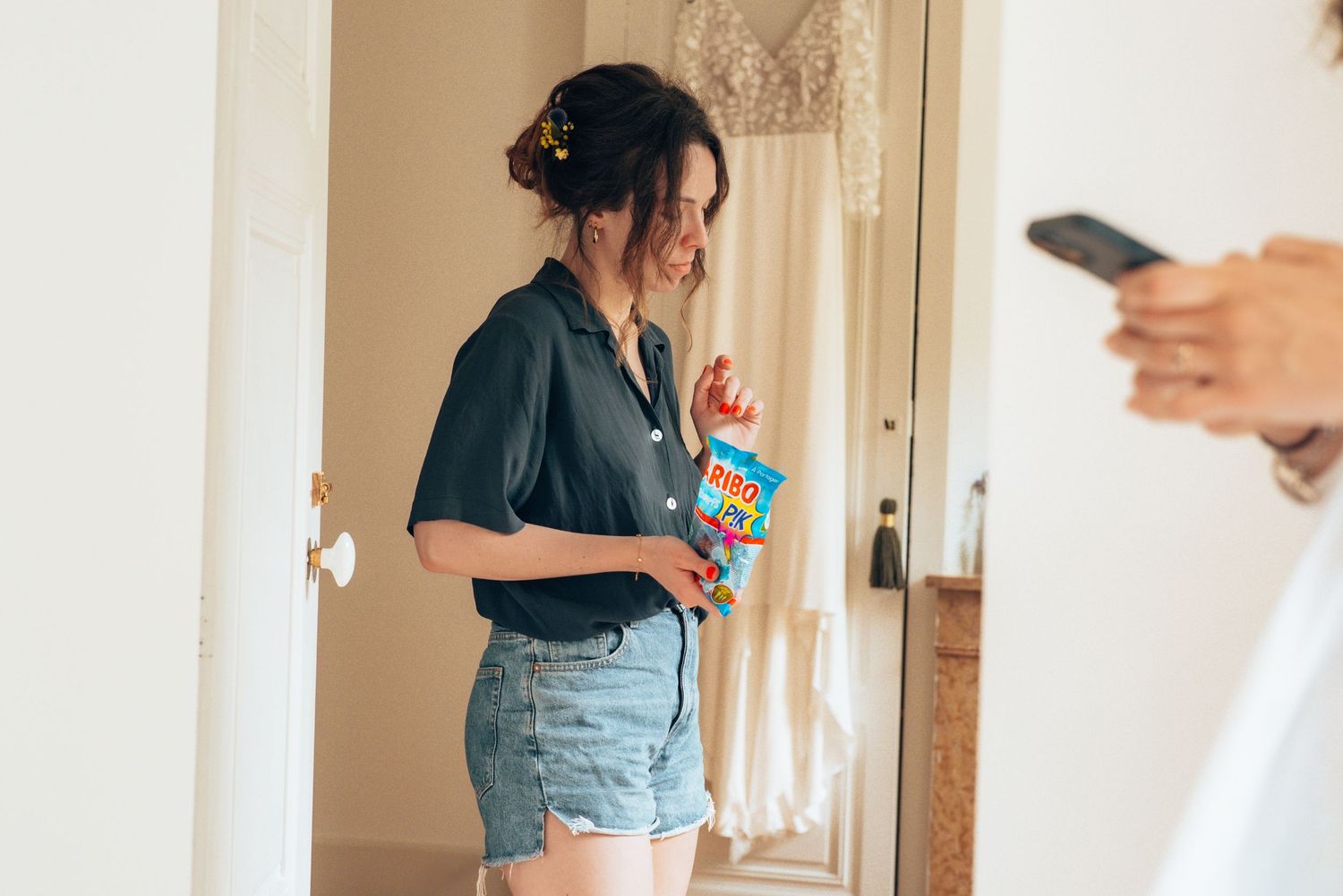 A person in denim shorts and navy shirt stands in a doorway while another person holds a phone in the foreground.