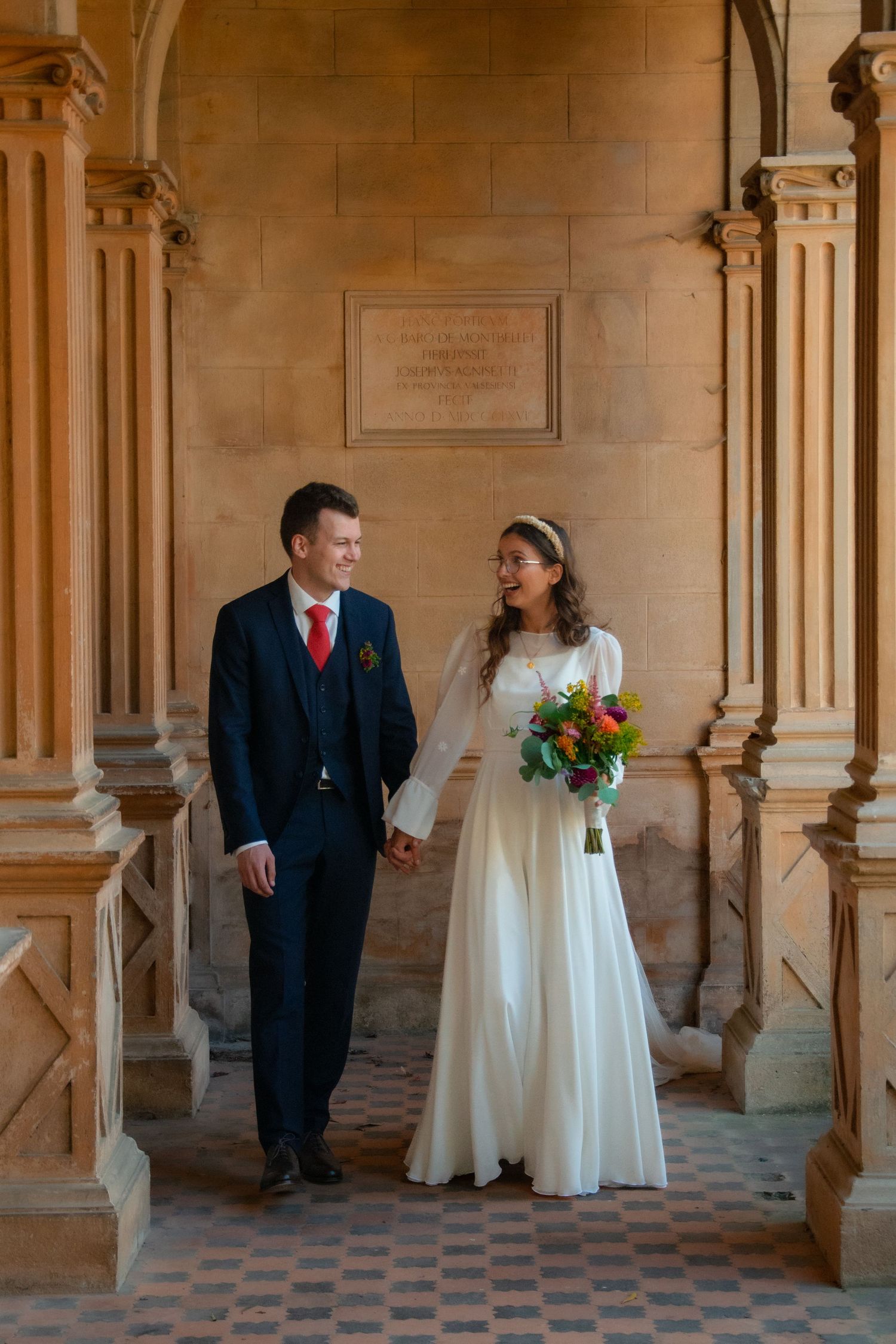 A couple in wedding attire holding hands while walking through an ornate stone corridor.