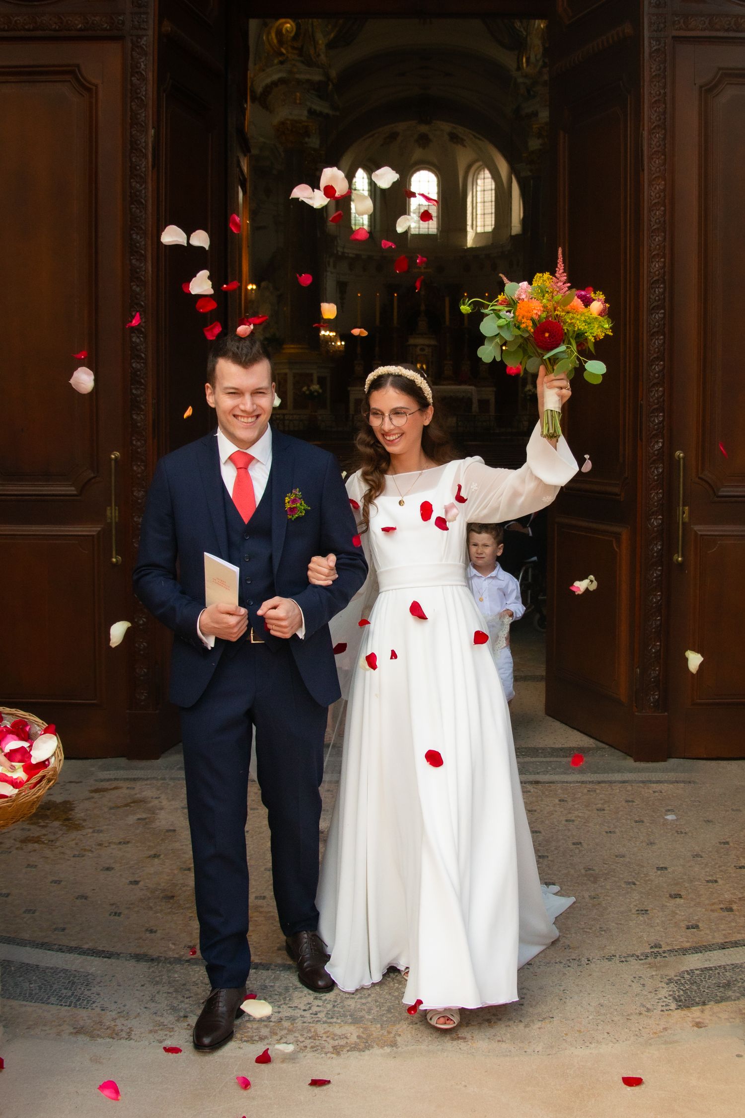 Newlyweds exit church under shower of rose petals, bride holding colorful bouquet.