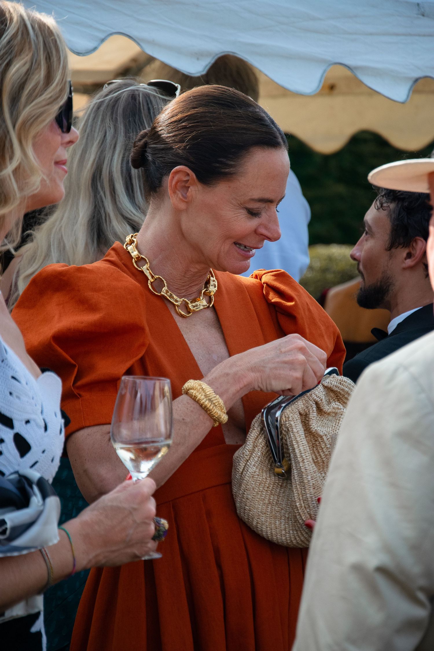 A group socializing at an outdoor event with someone wearing a striking orange dress and bold gold chain necklace.