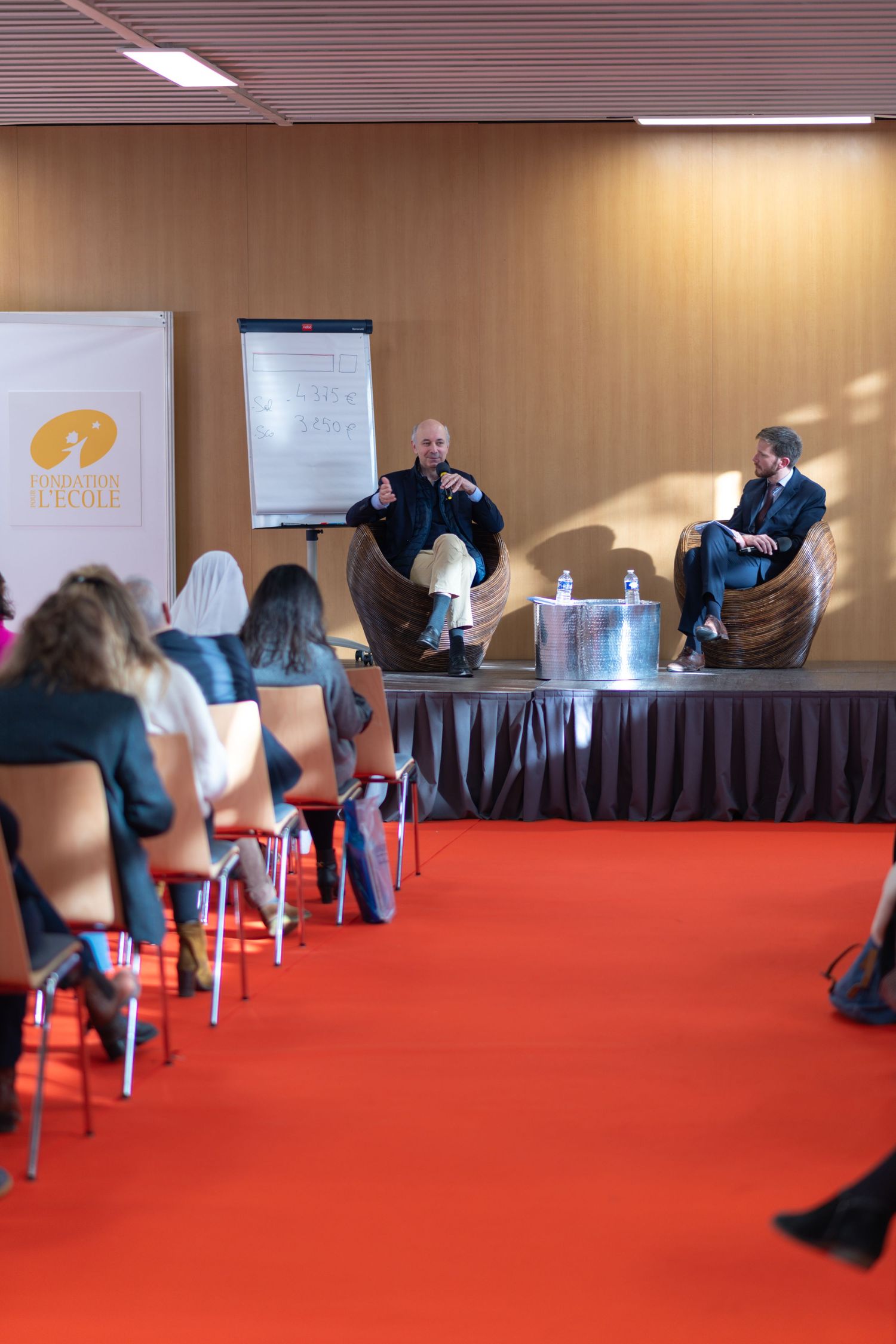 Panel discussion at a conference with speakers and audience seated on red carpet in a meeting room with beige walls.