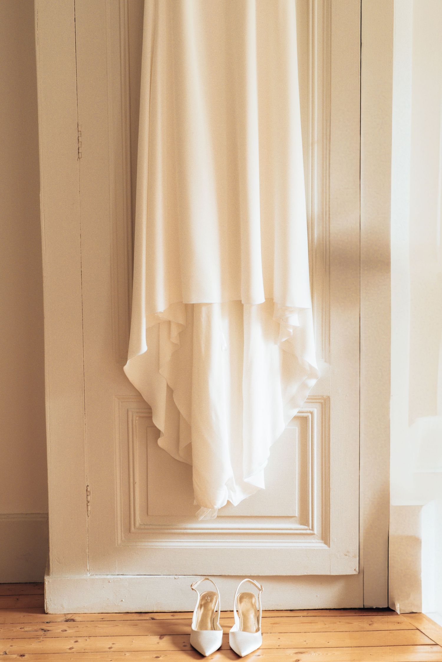 White wedding dress elegantly draped over a white door panel with ivory heels placed beneath on wooden flooring.