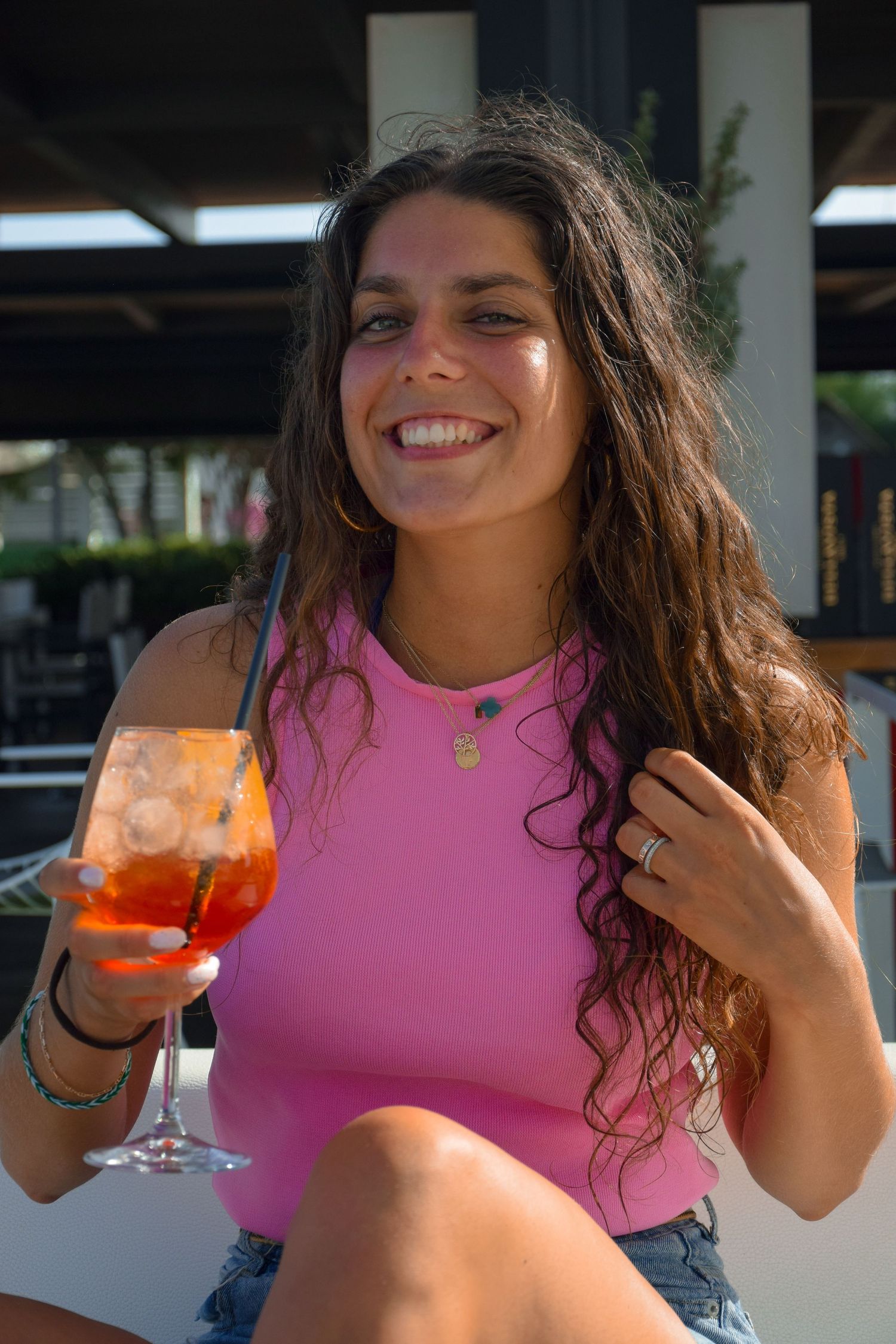 A person in a pink tank top smiles while holding an orange-colored cocktail at an outdoor restaurant.