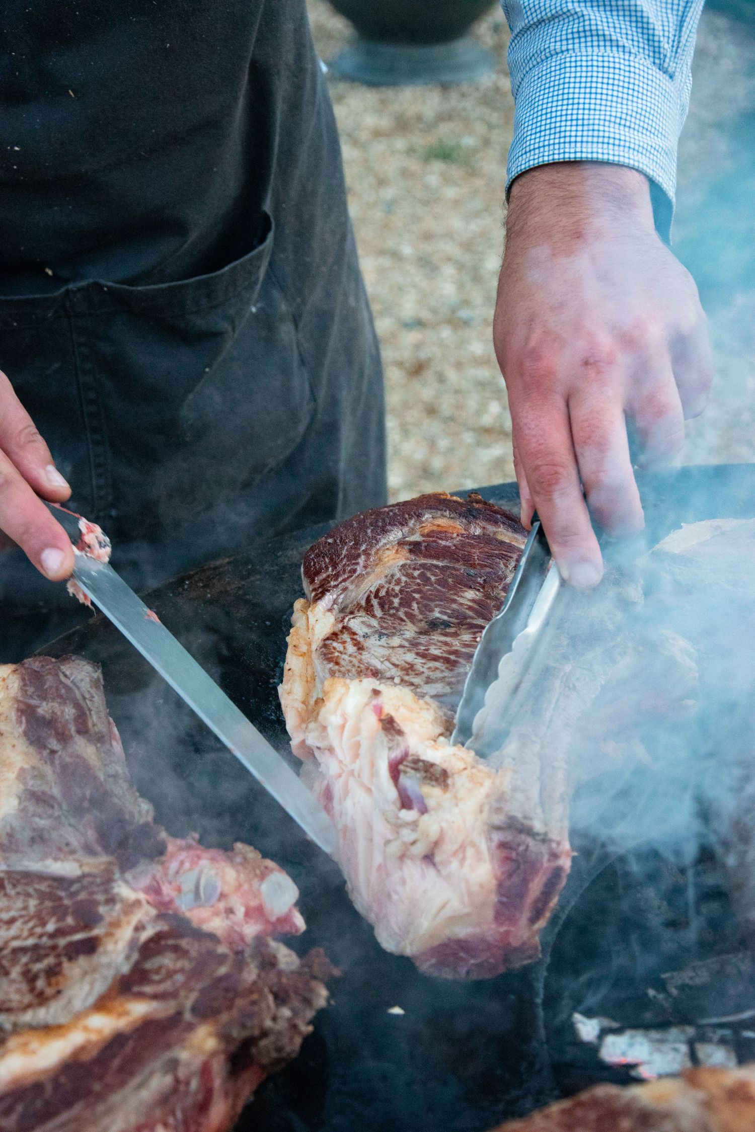 Hands slicing a large cut of grilled meat surrounded by smoke on an outdoor grill.