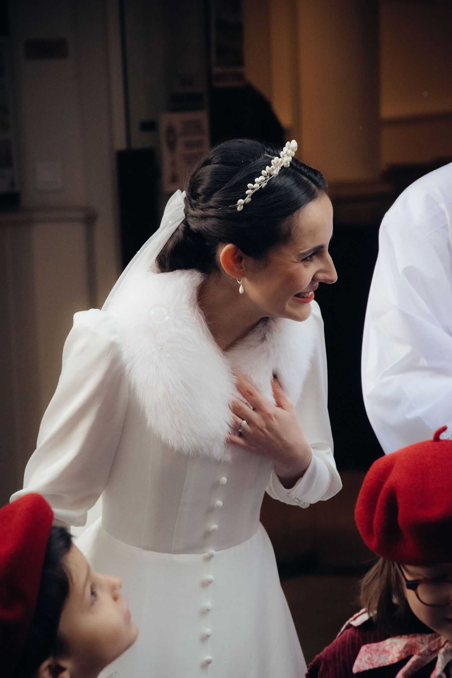 Elegant bride in white dress with fur collar and crystal tiara smiles while interacting with guests in red berets.