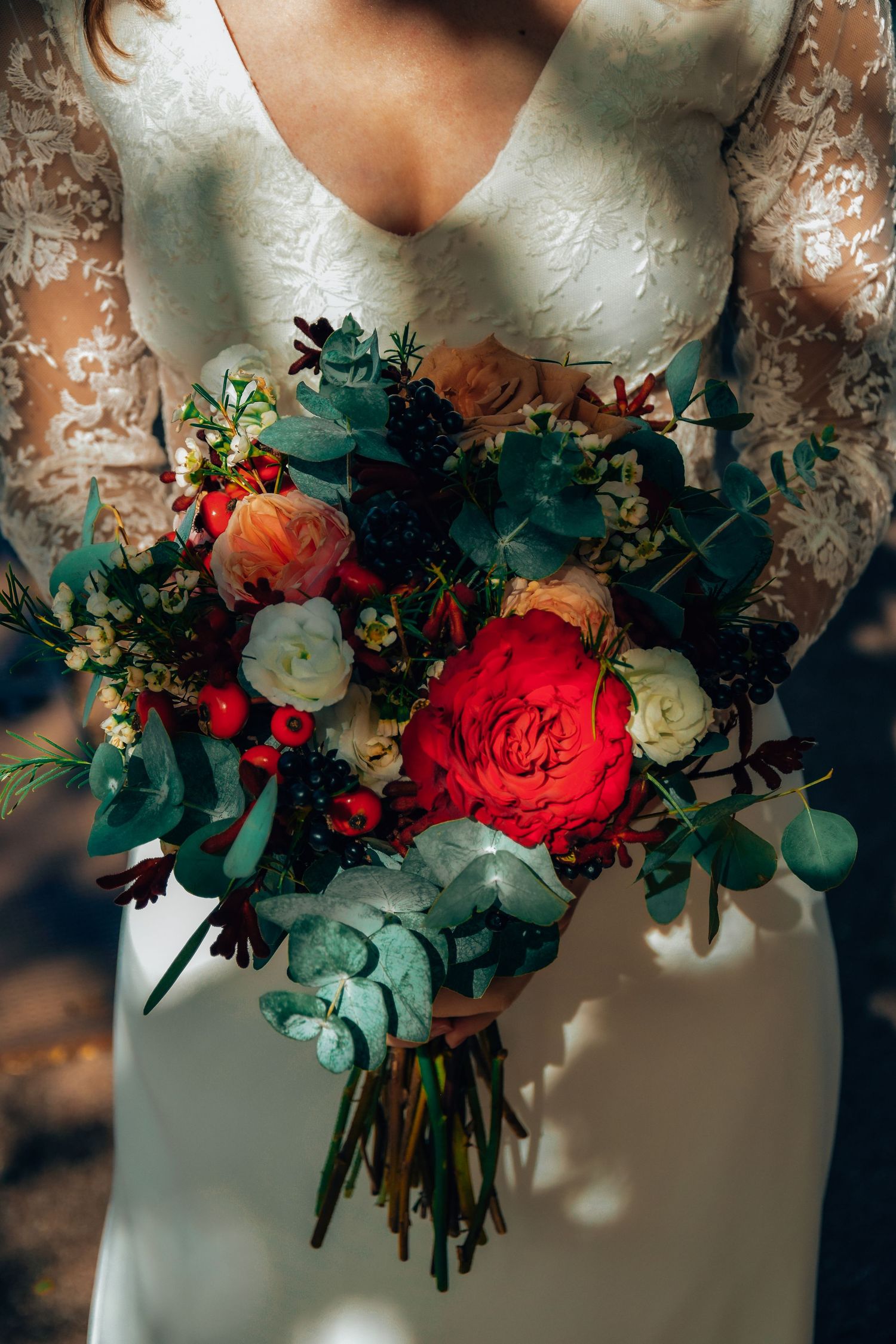 Vibrant bridal bouquet featuring red roses, white flowers and green eucalyptus against a lace wedding dress.