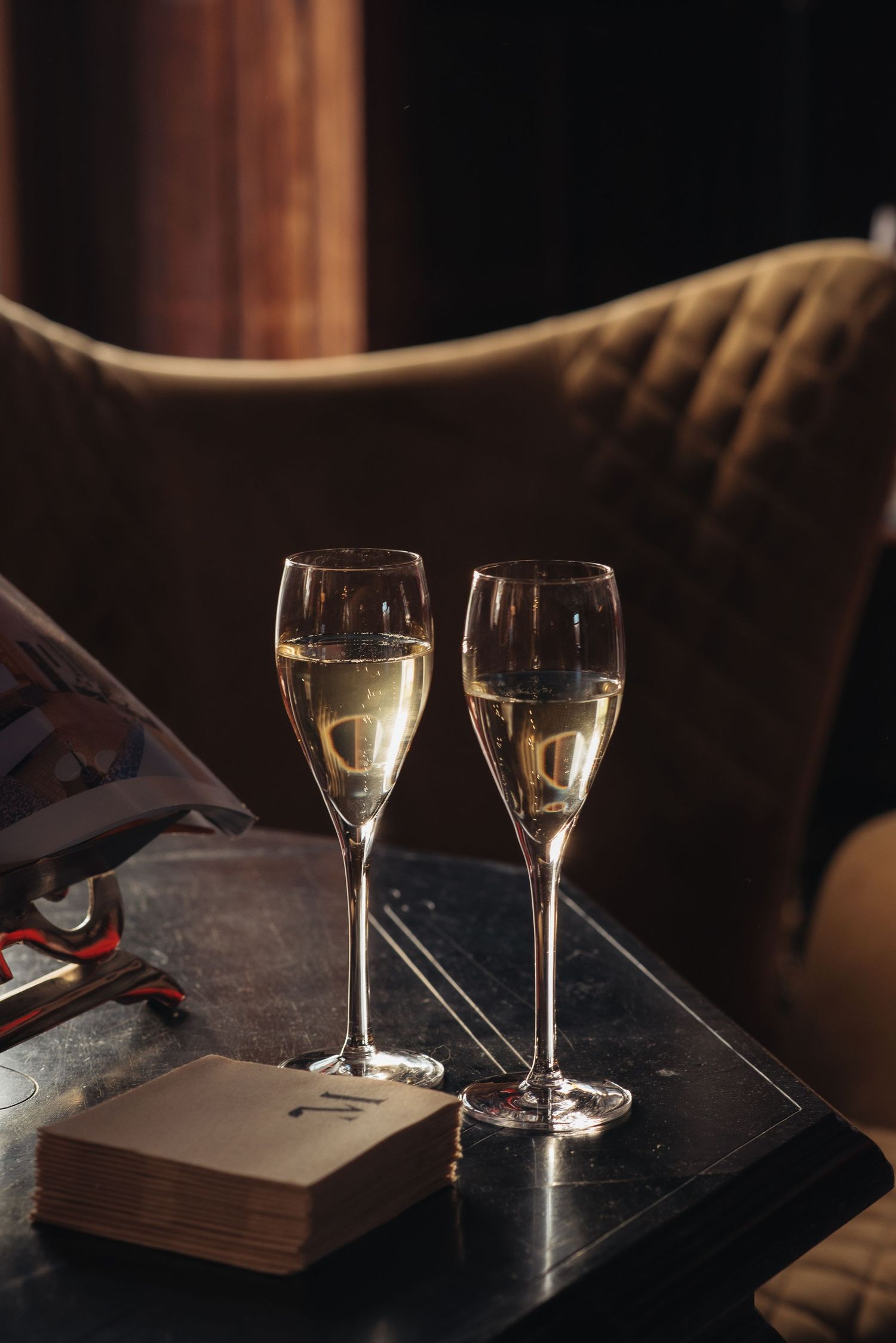 Two champagne flutes filled with sparkling wine sit on a dark table beside a book in a cozy setting.