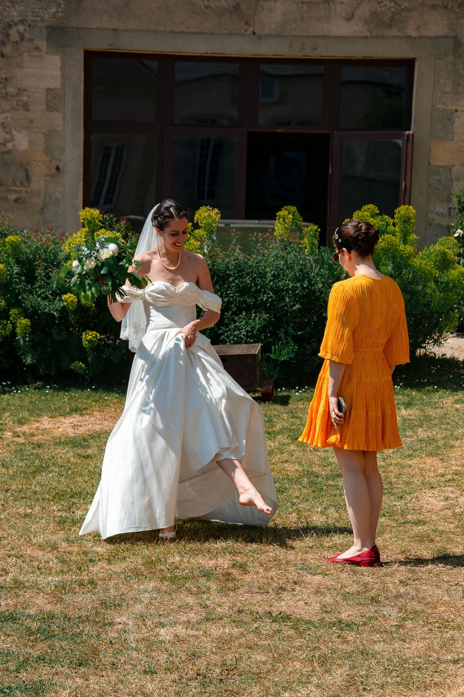 Two figures in contrasting white and orange attire interact playfully on dry grass near a stone building.