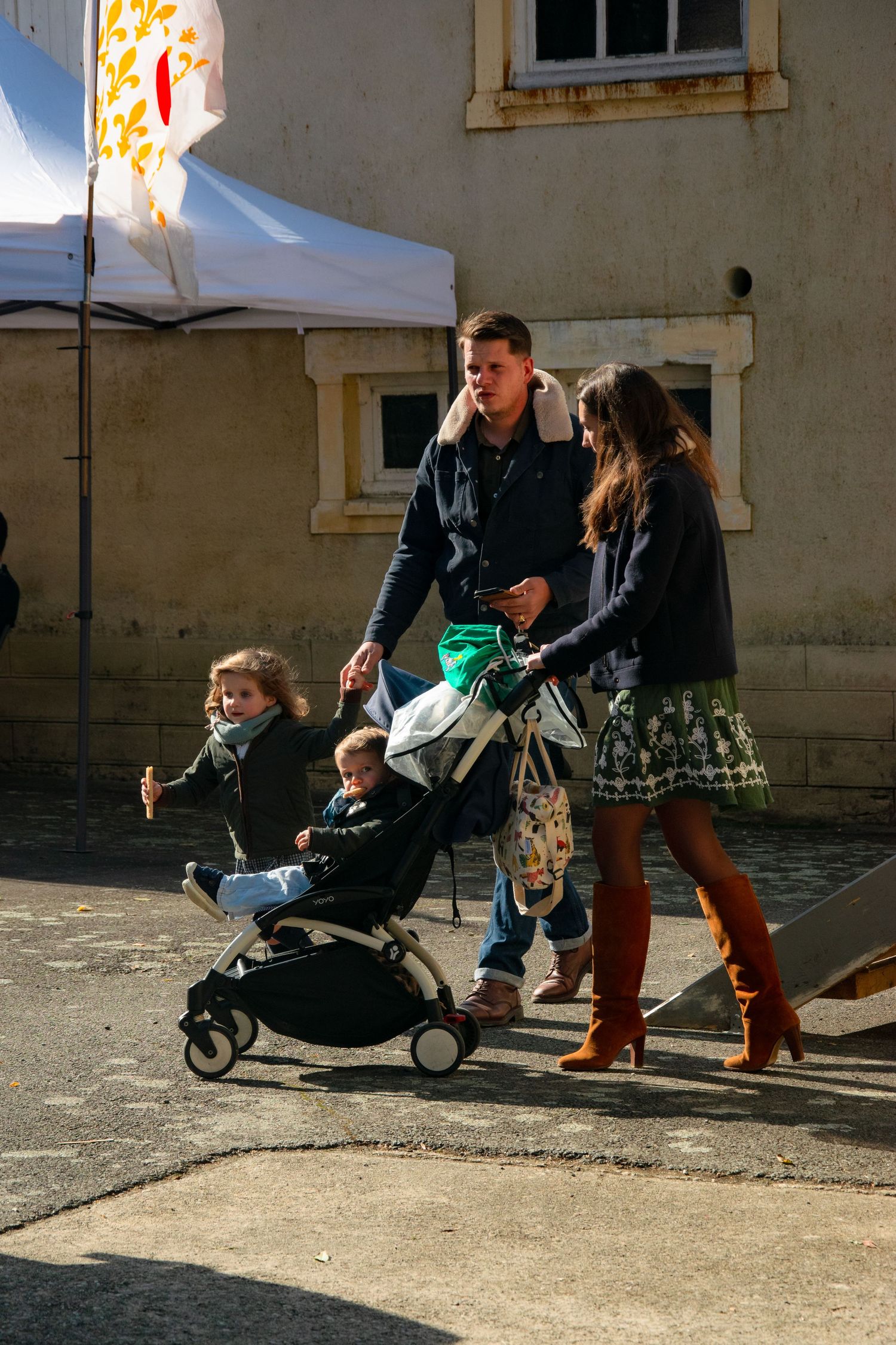 A couple pushes a stroller along a cobblestone street near a white tent and building.