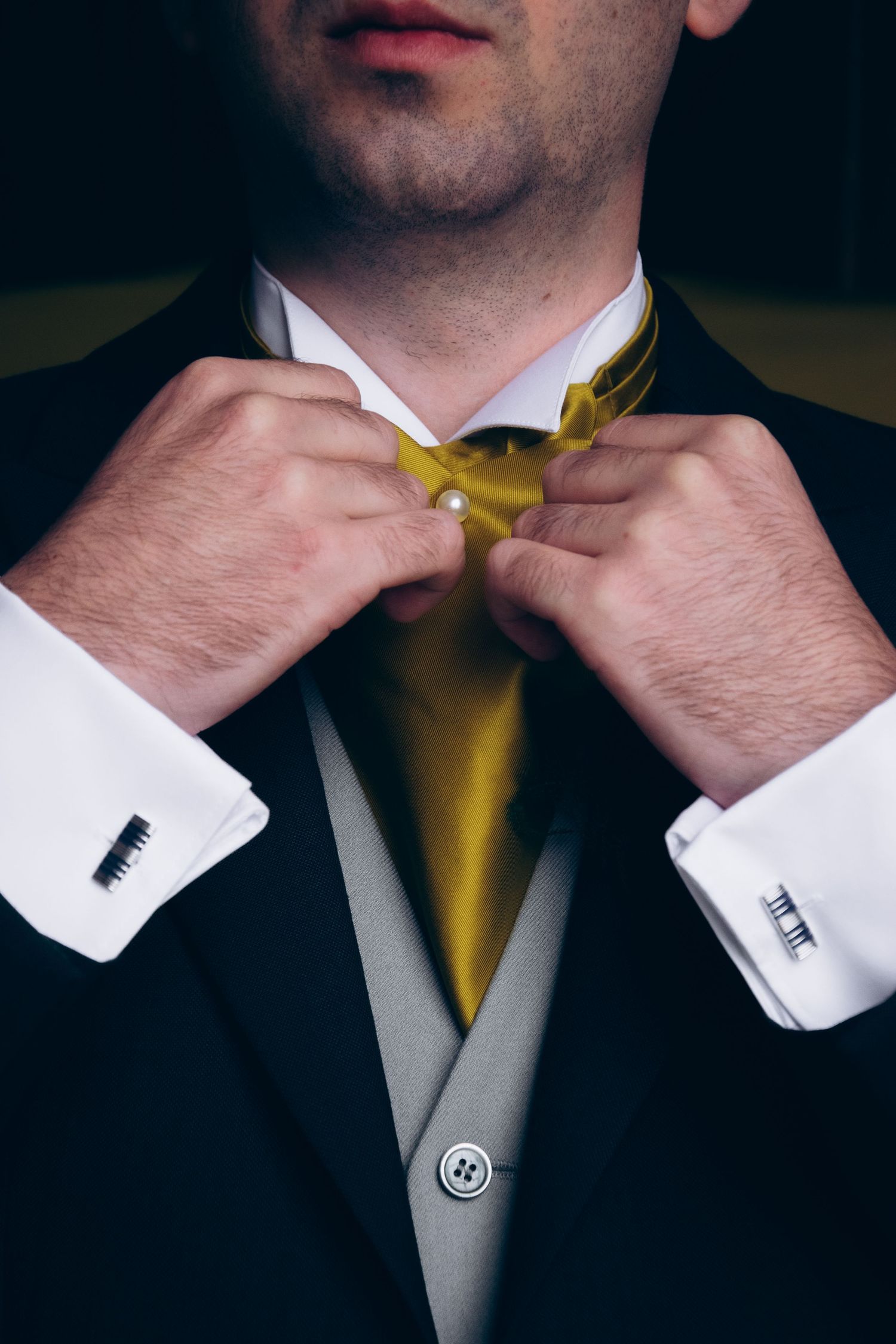 Close-up of groom adjusting gold tie with white cuffs and black suit jacket.