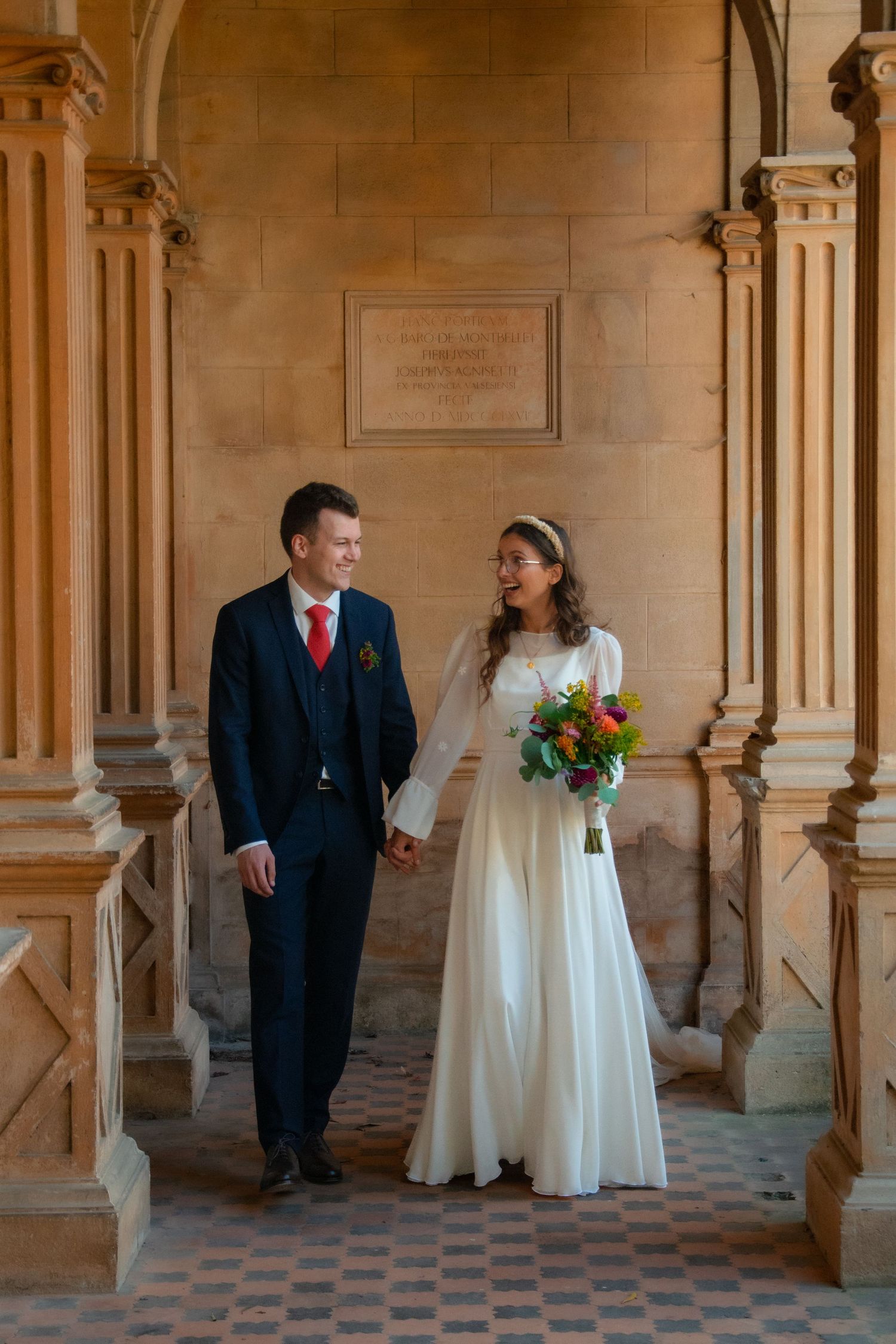 A wedding couple poses under ornate stone archways in a historic building.
