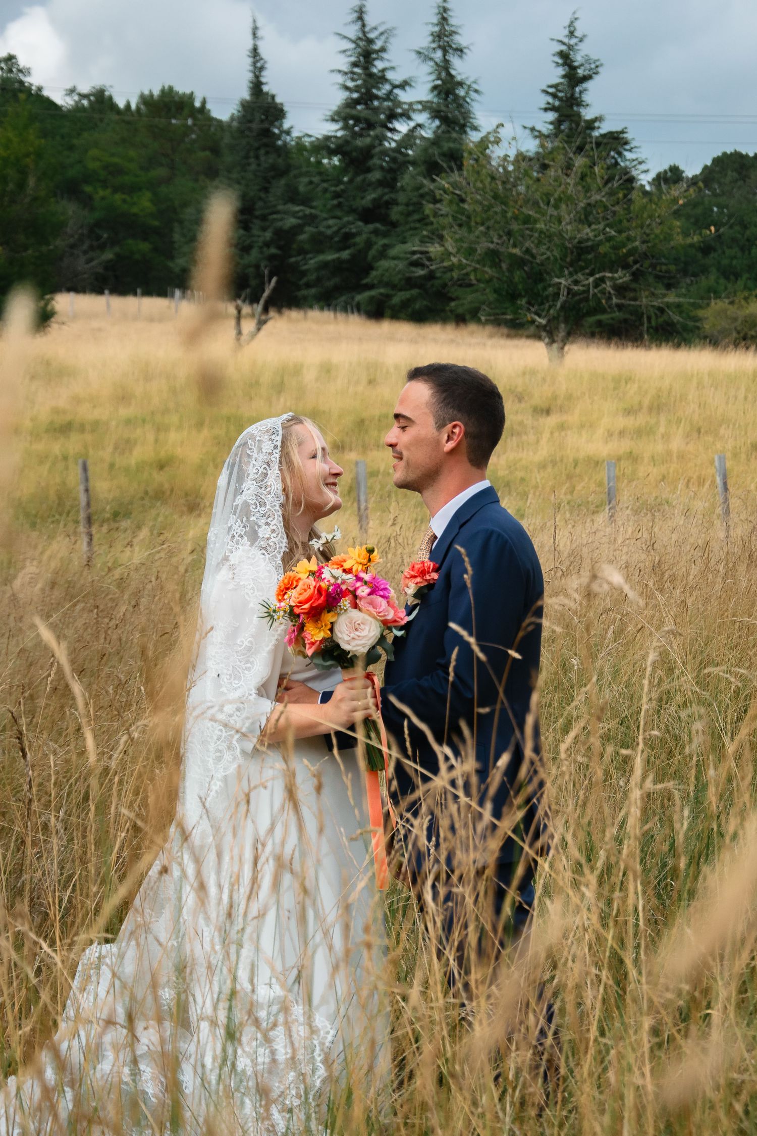 Newlywed couple shares intimate moment standing among tall grass and flowers at sunset in scenic outdoor setting.