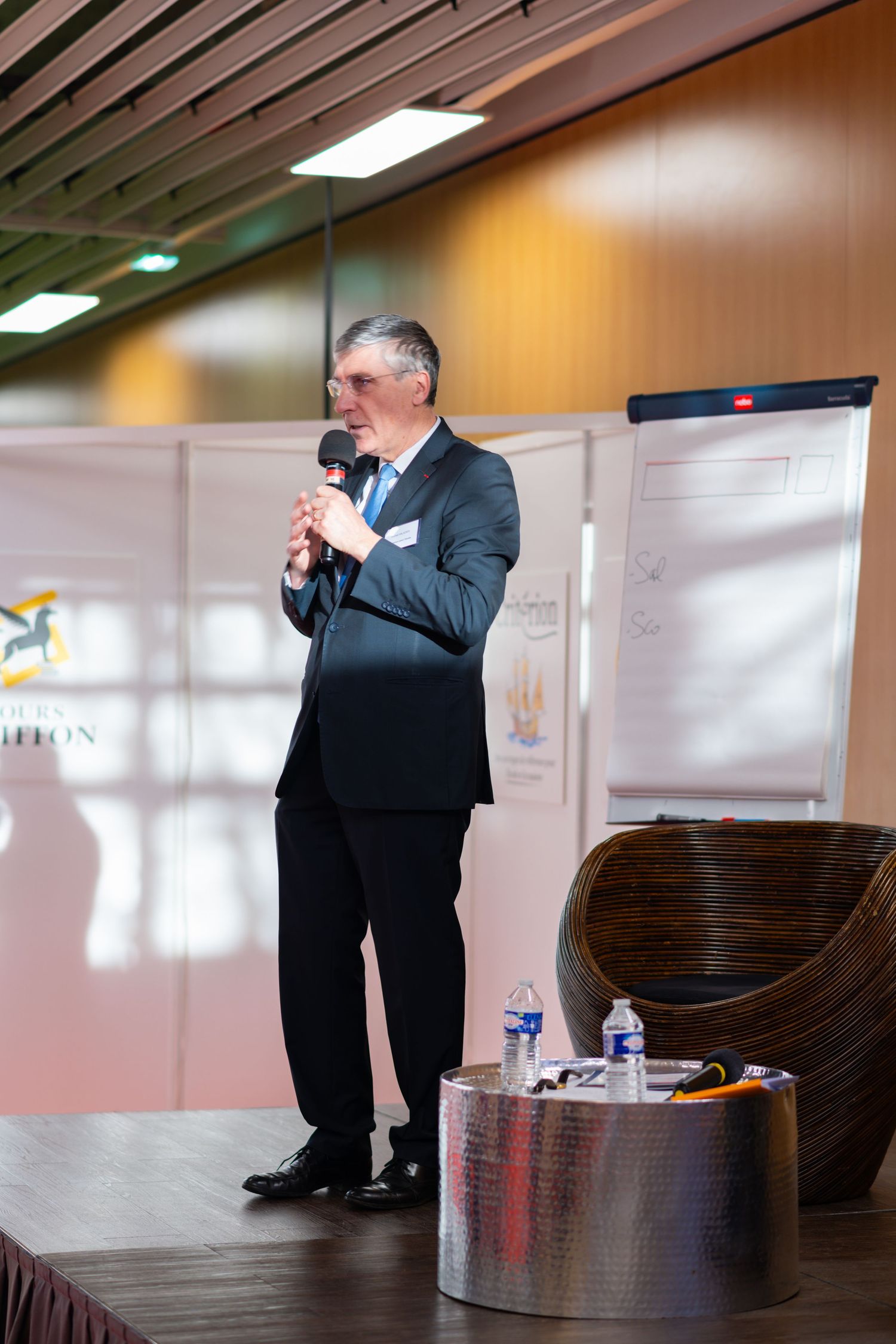 A person in formal business attire gestures while speaking during an indoor presentation event.