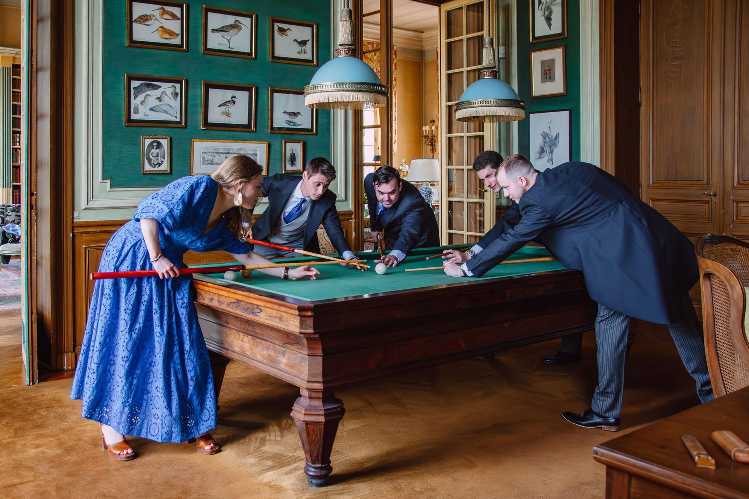 People in period costumes gather around an antique billiards table in a room with green walls and framed artwork.