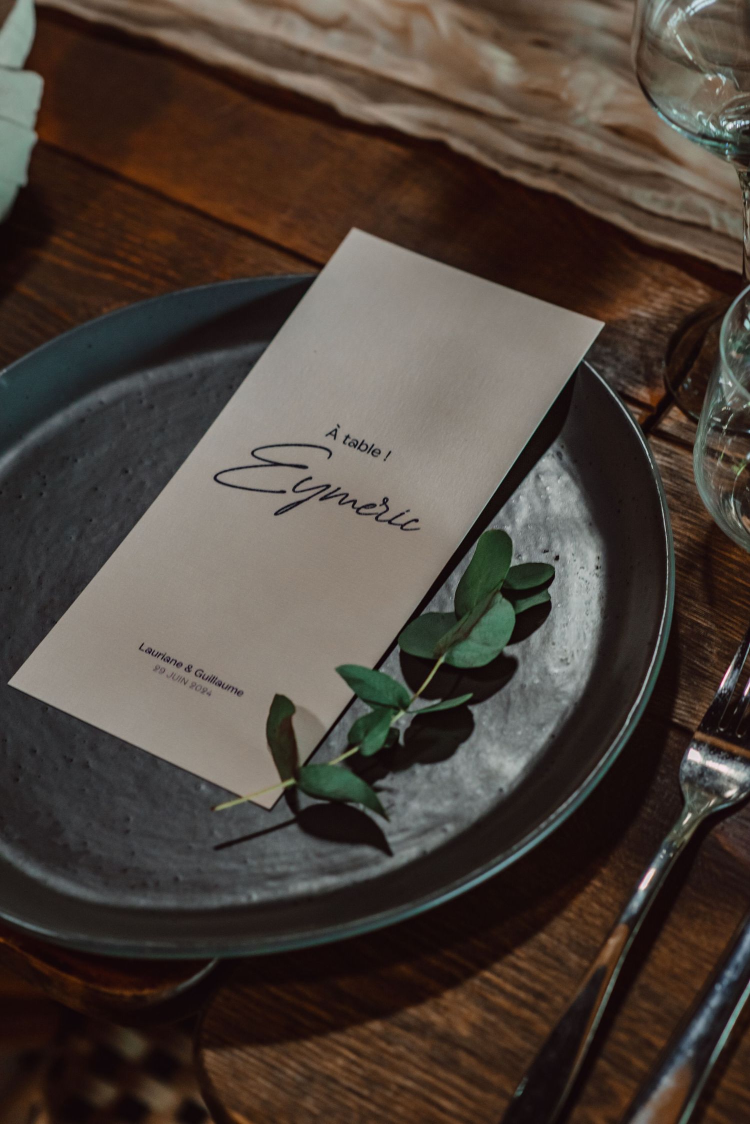 Elegant table setting with white menu card on dark plate accented with fresh eucalyptus sprig on wooden surface.