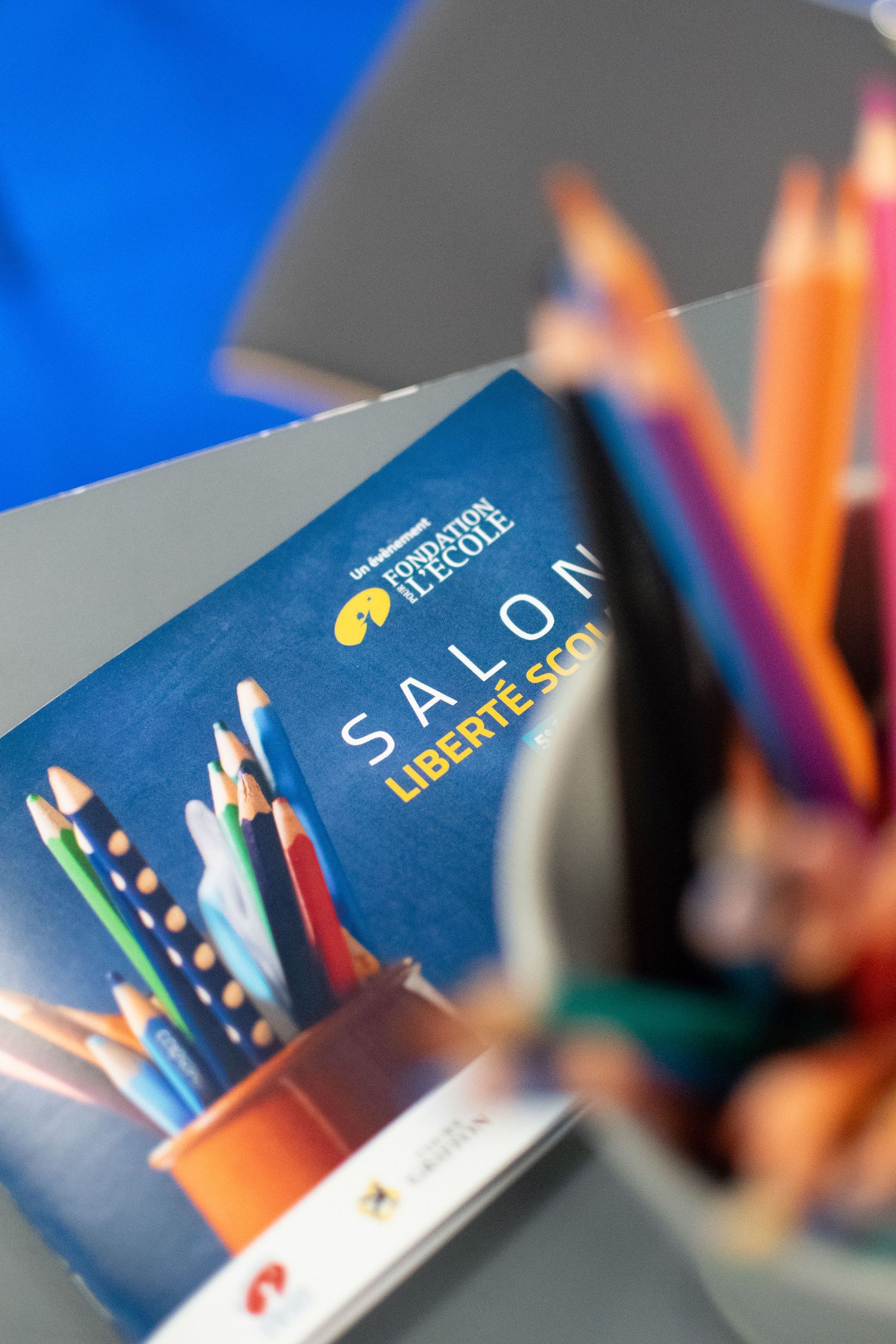 Colorful pencils arranged against a blue background with a blurred book spine or cover showing partial text.