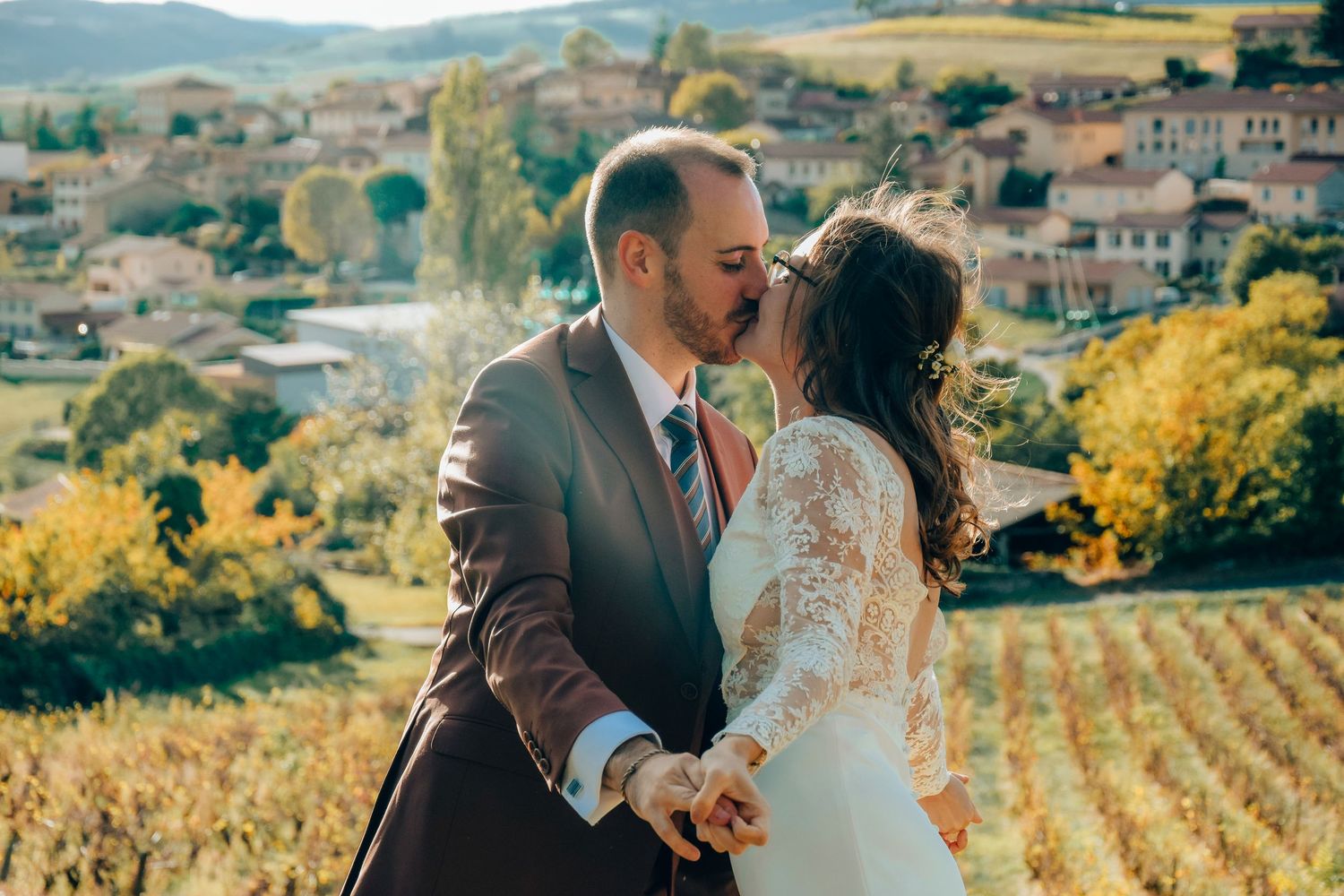 A couple embraces in a romantic vineyard wedding photo with rolling hills and a rustic village in the background.