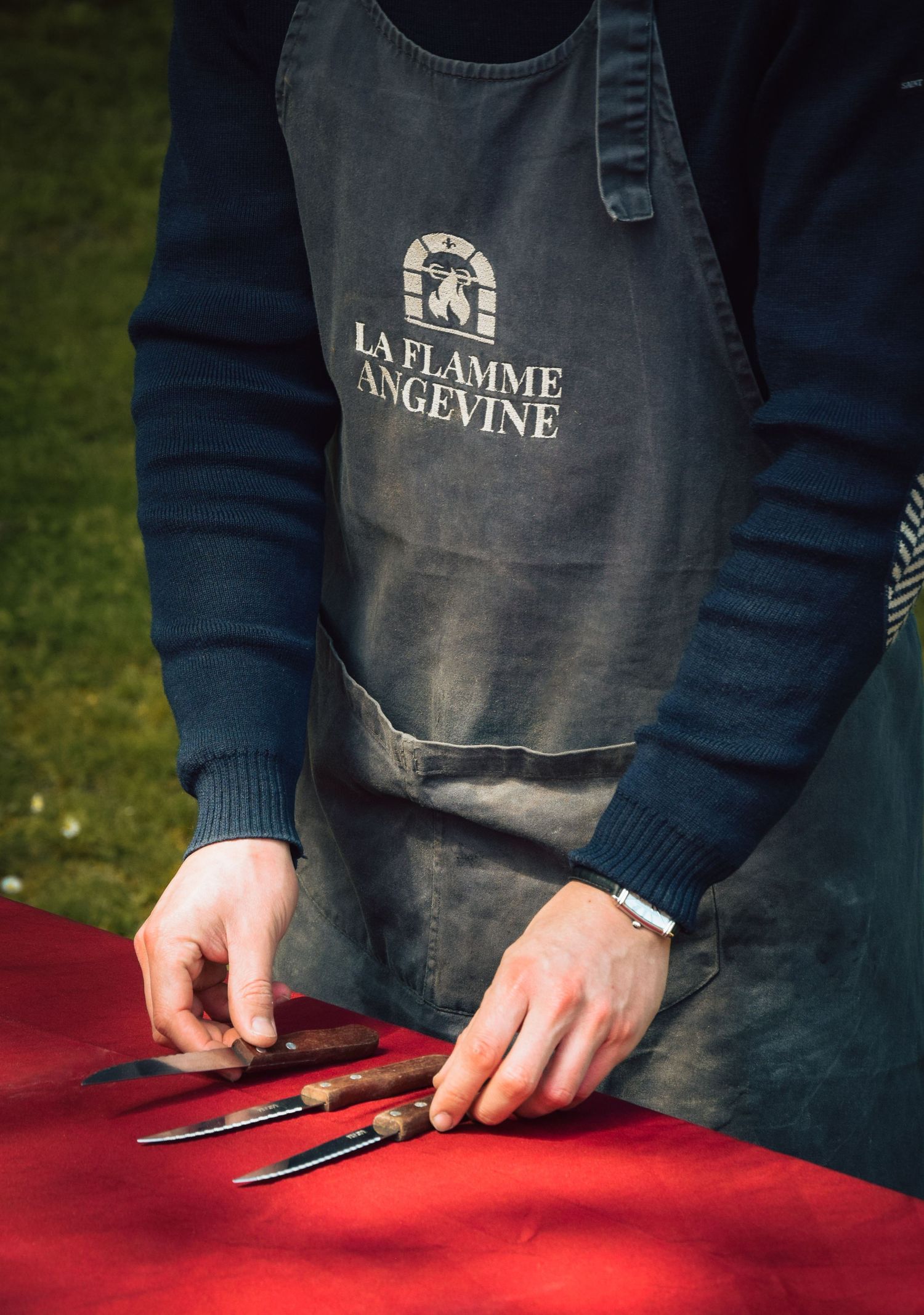 Close-up of hands wearing a La Flamme Angevine apron preparing utensils on a red tablecloth.