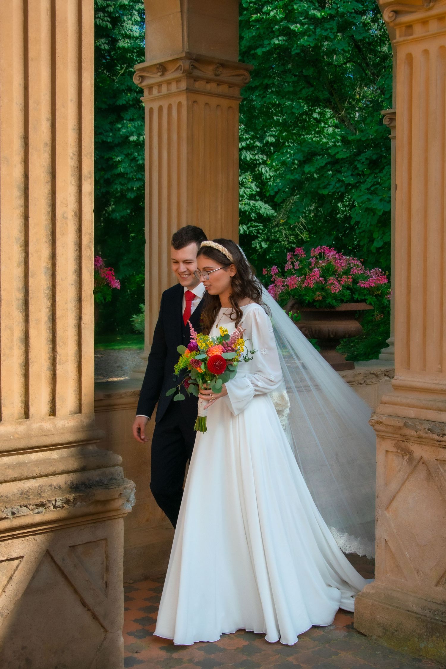 Wedding couple walking between elegant stone columns surrounded by lush greenery and pink flowers.