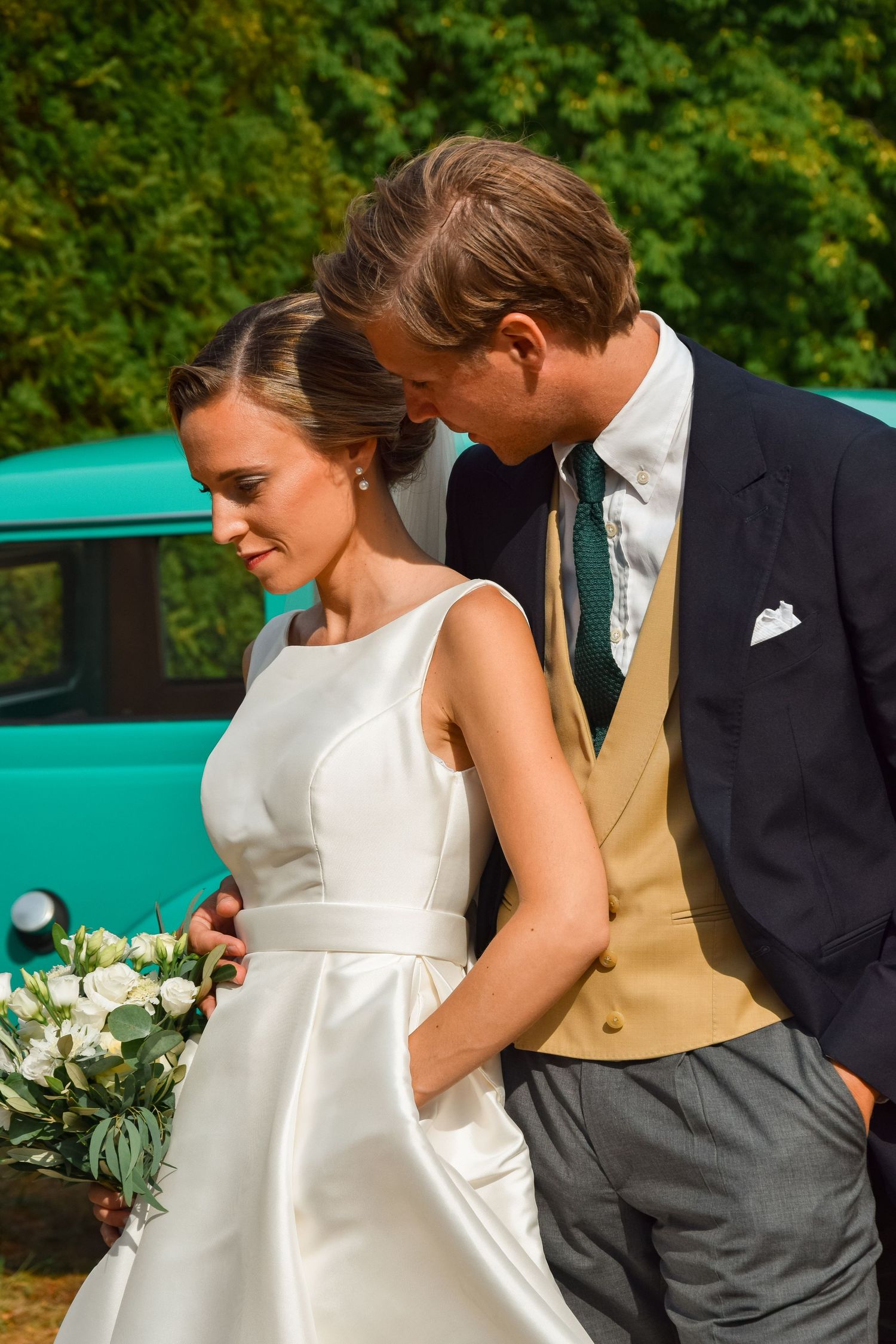 A couple on their wedding day poses outdoors with a classic turquoise car in the background and white floral bouquet.