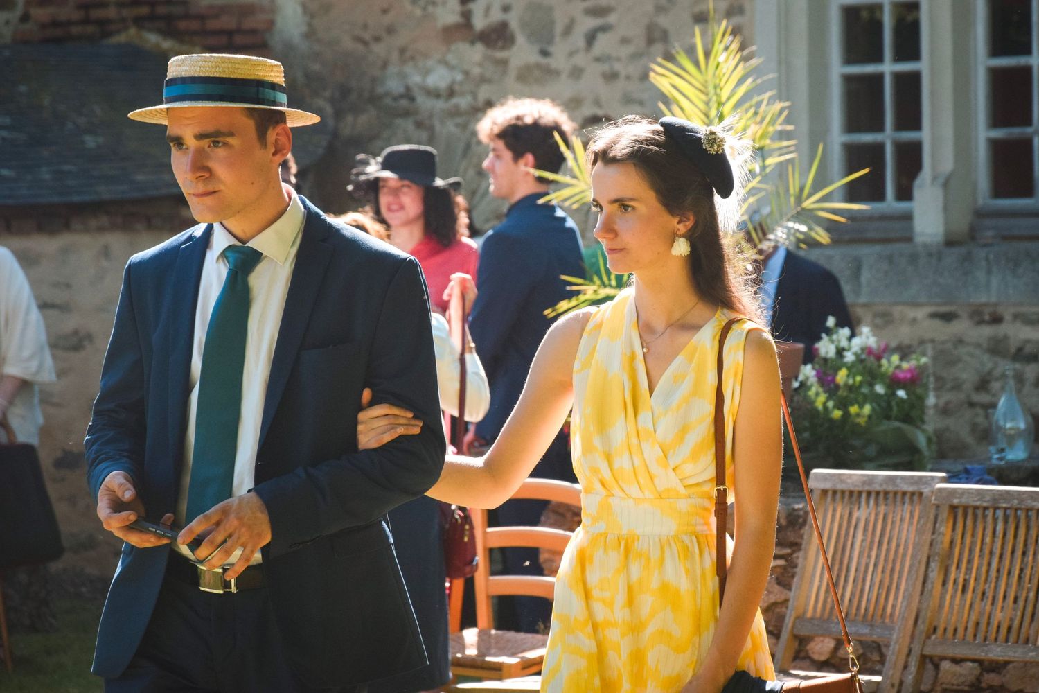 Couple in vintage attire walking outdoors, man in boater hat and woman in yellow sundress.