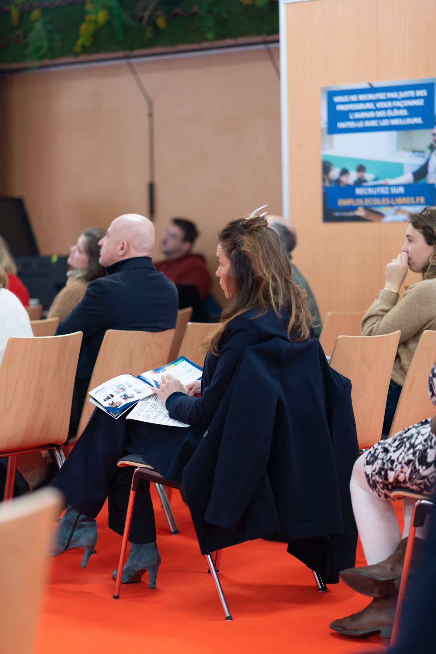 Conference attendees seated on orange carpet reviewing materials during presentation.