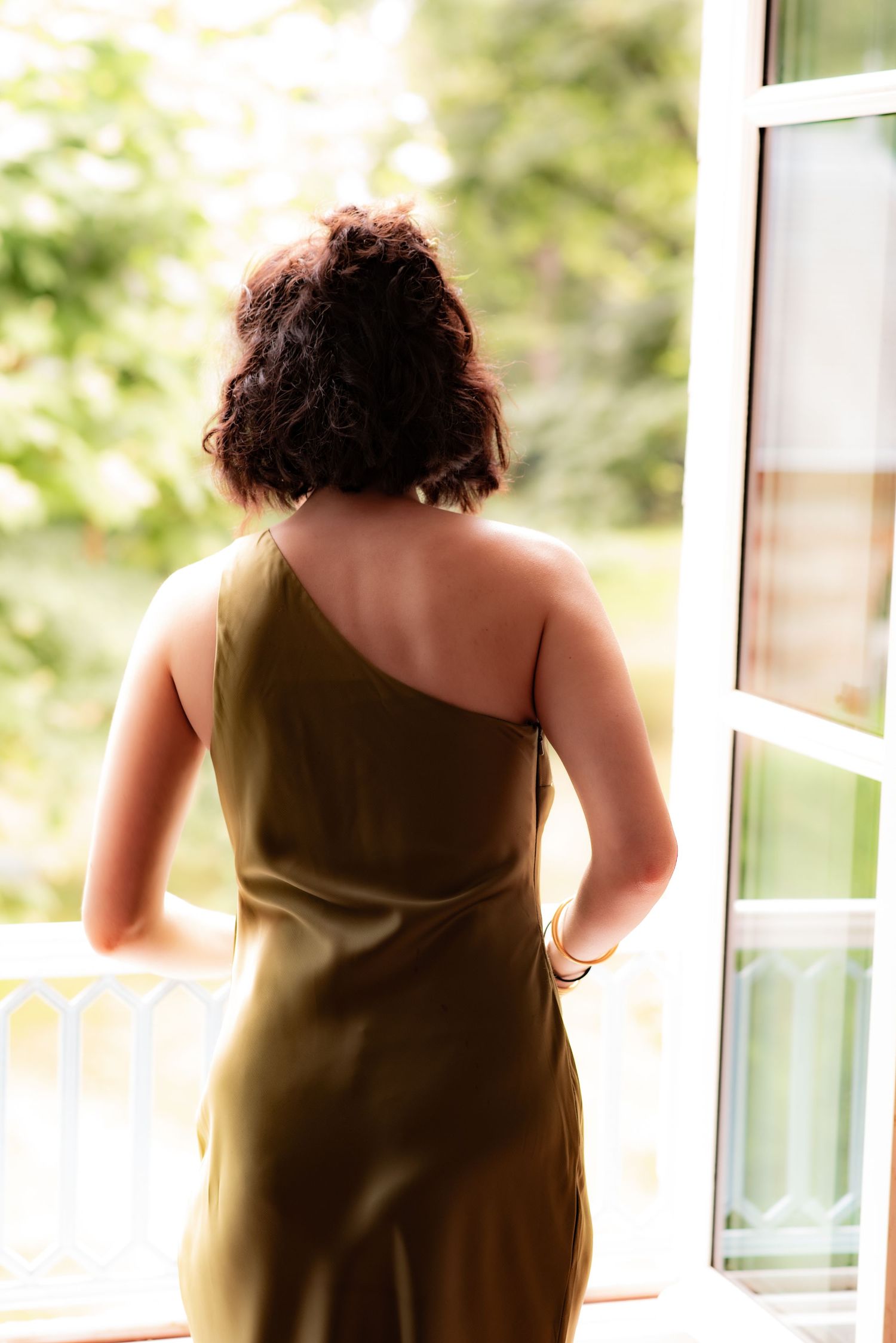 Back view of person in olive one-shoulder dress looking out through open French doors on bright sunny day.