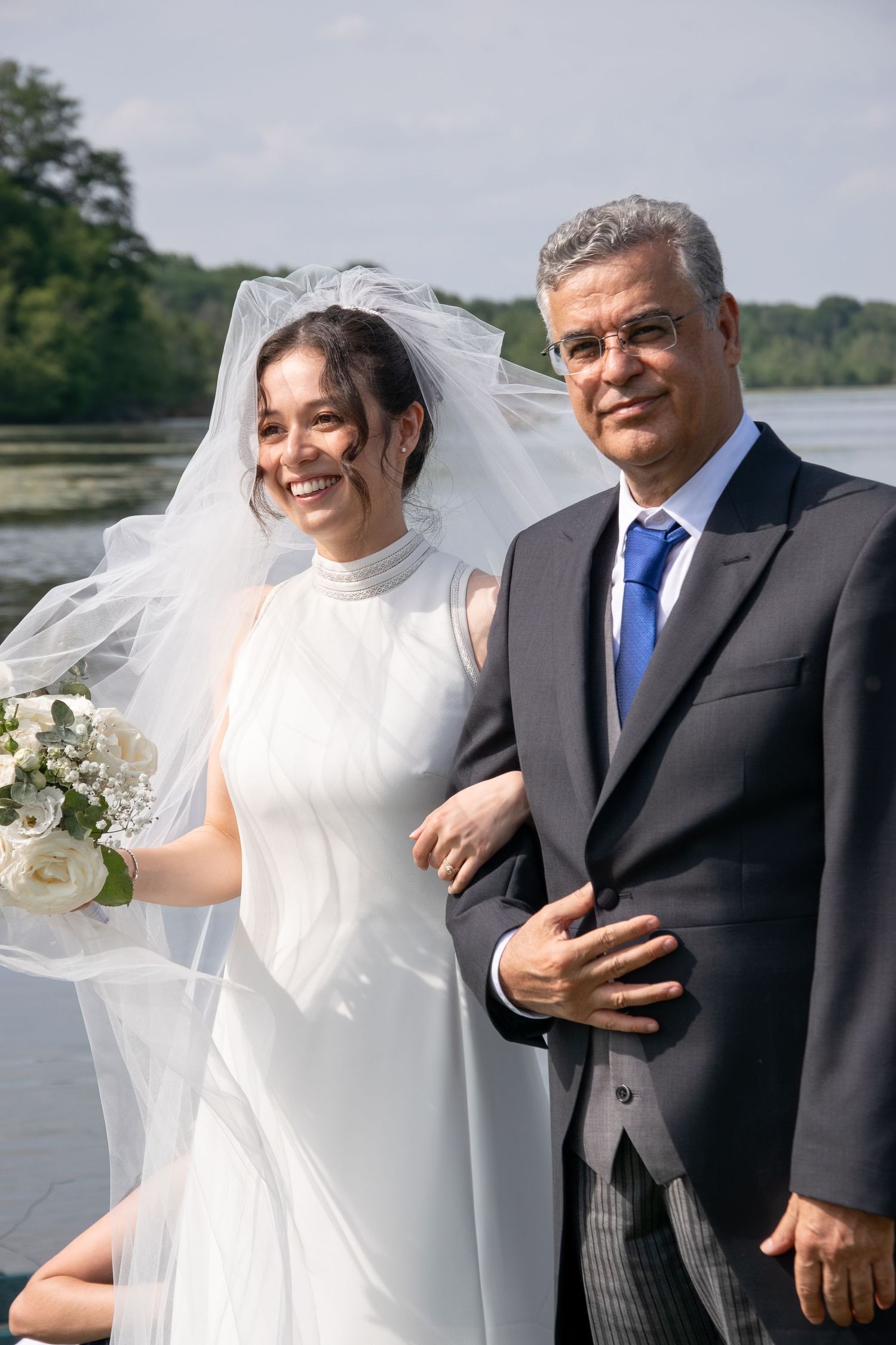 A bride and her escort share joyful moments by the waterside during an outdoor wedding ceremony.
