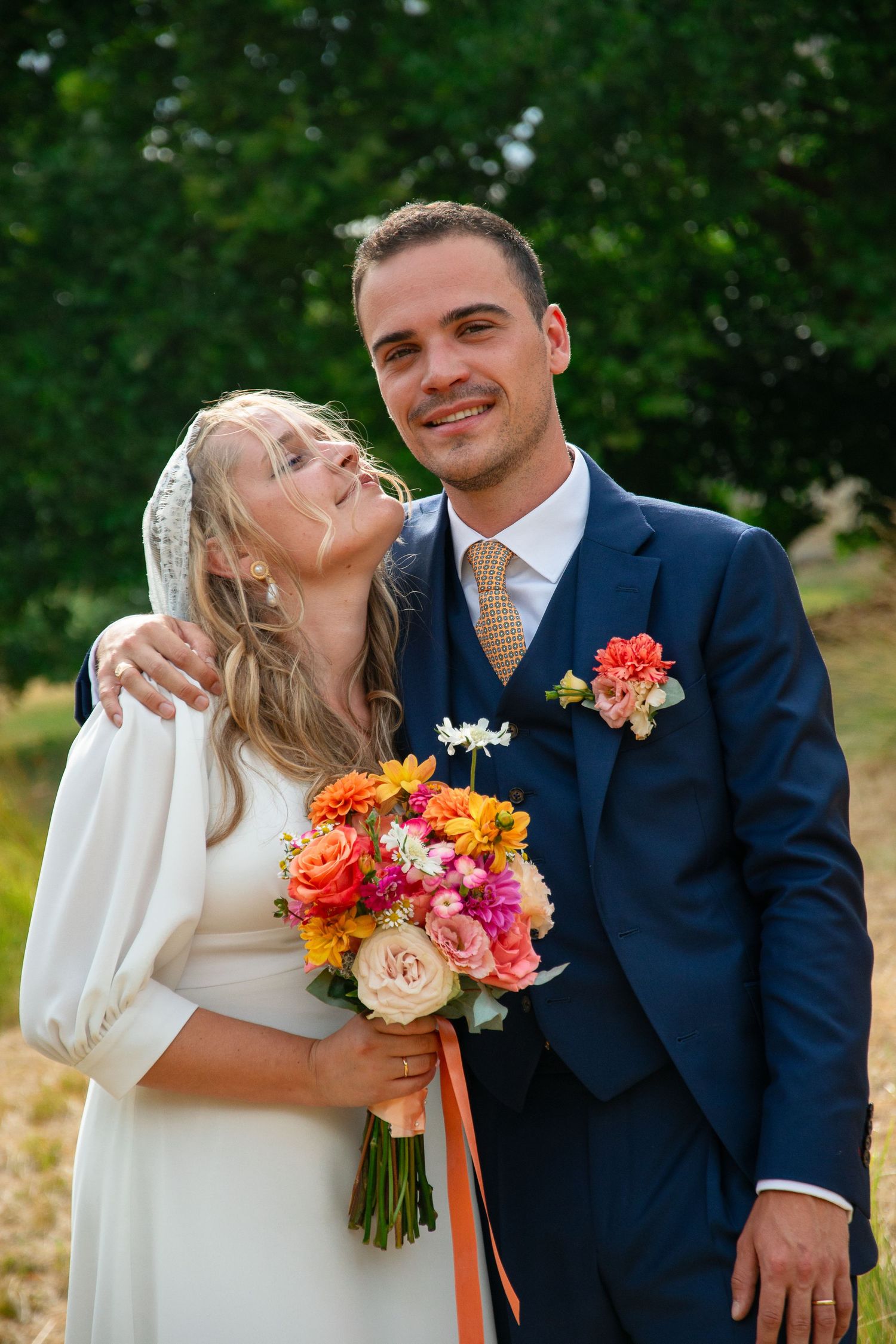 A bride and groom in a navy suit share romantic moments outdoors with a colorful orange and pink bridal bouquet.