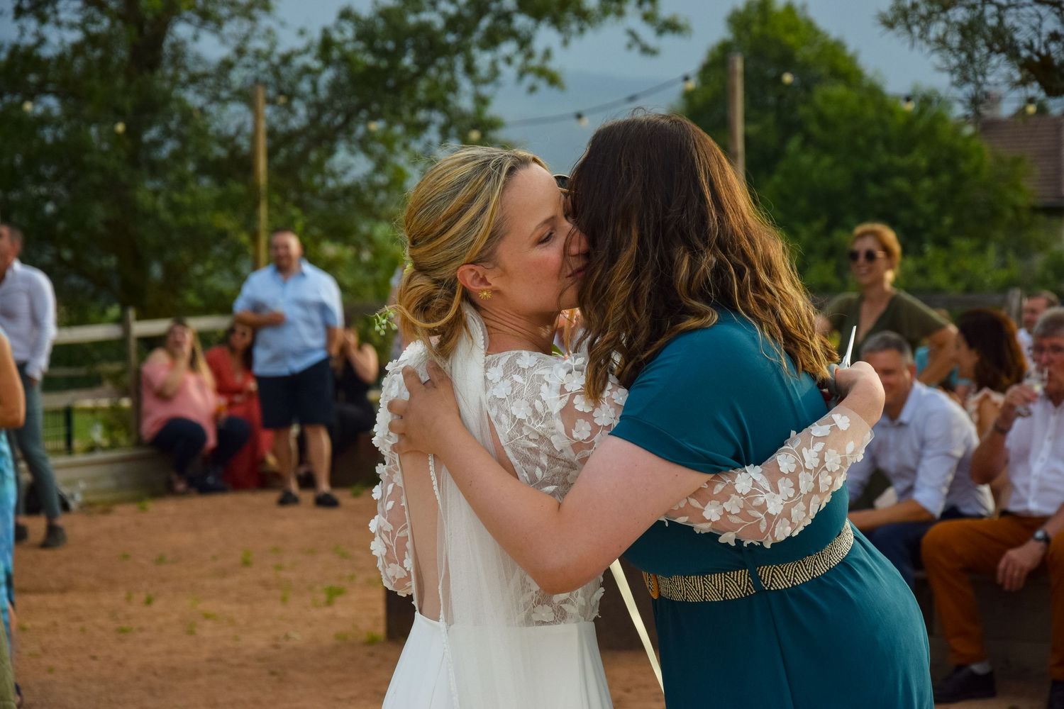 Two people sharing an emotional embrace at an outdoor wedding celebration with guests seated in background.